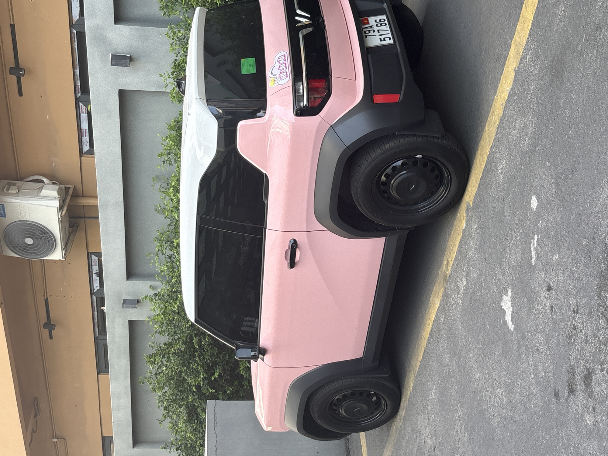 A pink, two-door electric vehicle is parked on asphalt in what appears to be an outdoor parking area. The car has black trim around the wheel wells and a white roof. A license plate with the characters "79A 517.86" is visible on the rear of the vehicle. There is a sticker on the rear passenger side of the car. In the background, there is a gray wall and some greenery, with a building and an air conditioning unit visible above the wall. The image was taken in Nha Trang Ward, Vietnam.