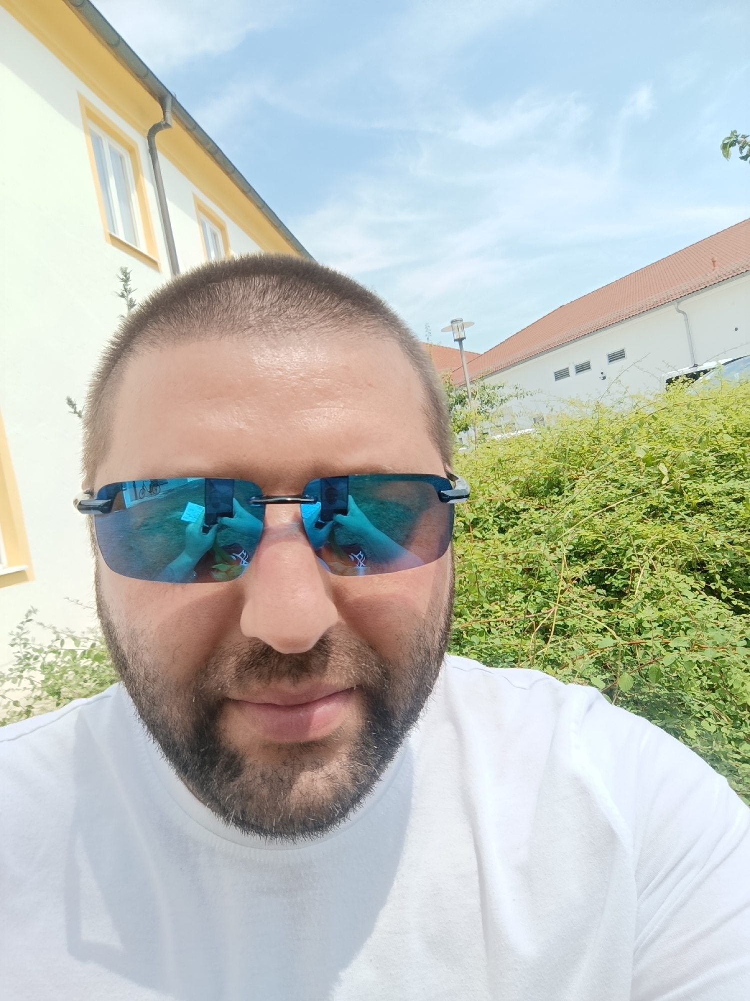 A close-up, selfie-style photograph features a man wearing a white t-shirt and blue-tinted sunglasses. The man has short, dark hair and a beard. The sunglasses reflect the environment, showing a person holding a phone and a bicycle. The background consists of a building with a yellow facade and a red-tiled roof, partially obscured by dense green foliage. The sky is blue with scattered clouds. The scene appears to be outdoors, likely in a residential or commercial area of Beilngries, Germany, given the architectural style of the building and the commonality of such settings in the region.