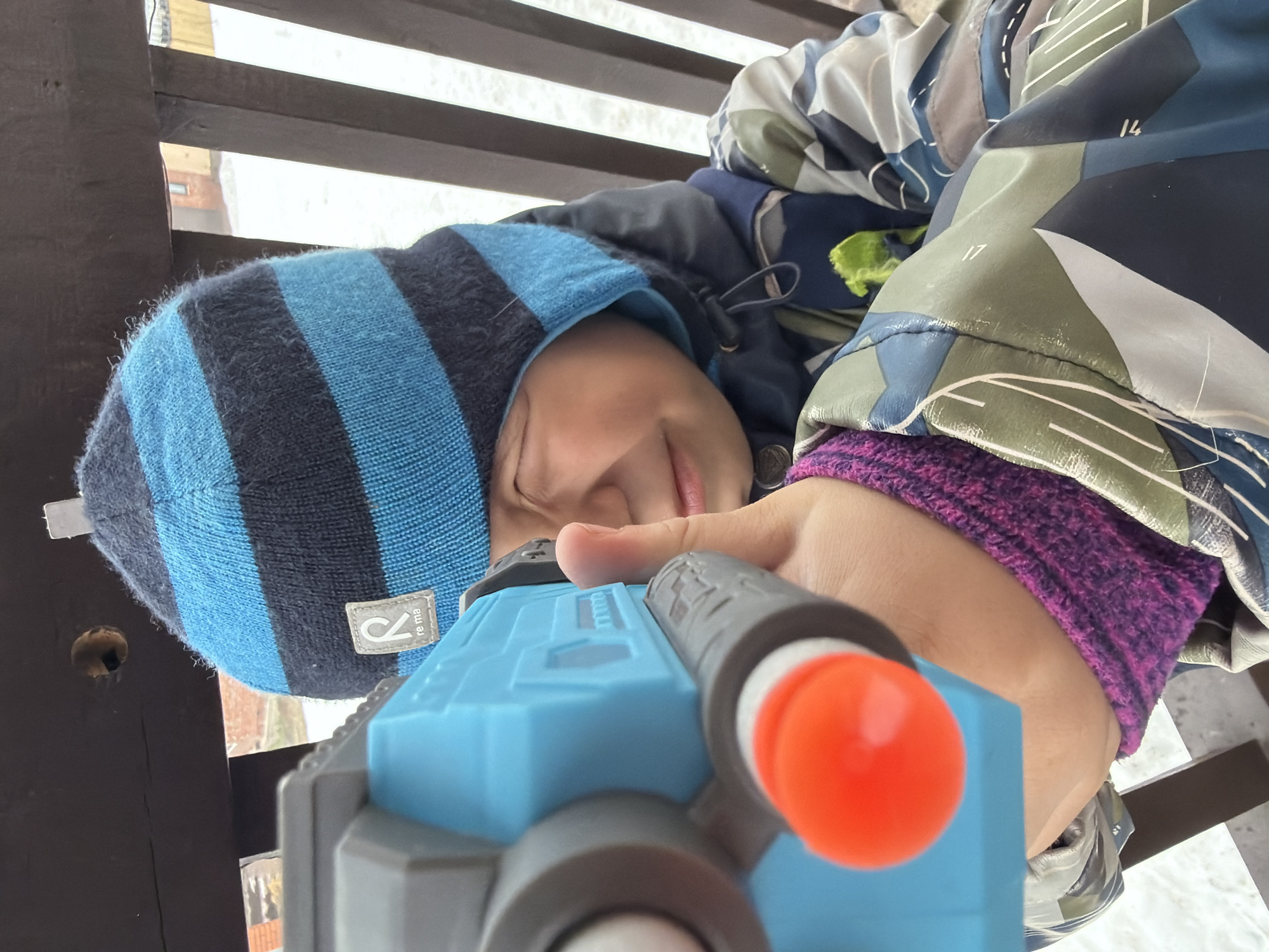 A child wearing a blue and black striped hat, a patterned jacket with purple cuffs, and holding a blue toy gun with an orange tip is lying on their side outdoors. The background consists of wooden slats, possibly a fence or structure, with snow visible through the gaps. The location is the city of Chaikovsky, Russia.