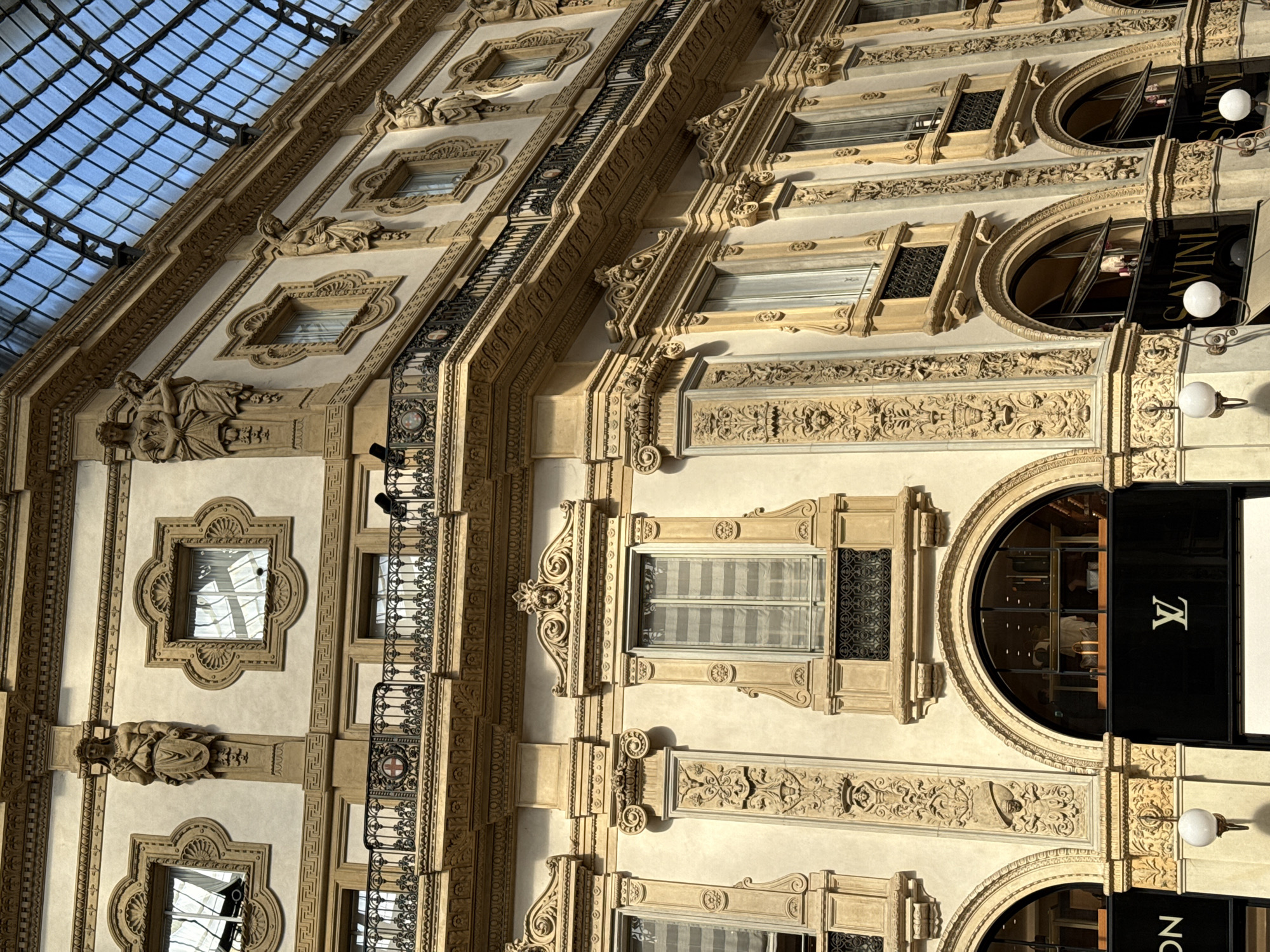 The image showcases an ornate architectural facade in Milan, Italy, likely within the Galleria Vittorio Emanuele II. The building features intricate carvings, classical statues, wrought iron balconies, and arched windows. One window is covered with a Louis Vuitton awning with the LV logo. The architecture suggests a historical structure, and the light in the image indicates daytime. The sky is visible through the glass roof structure.