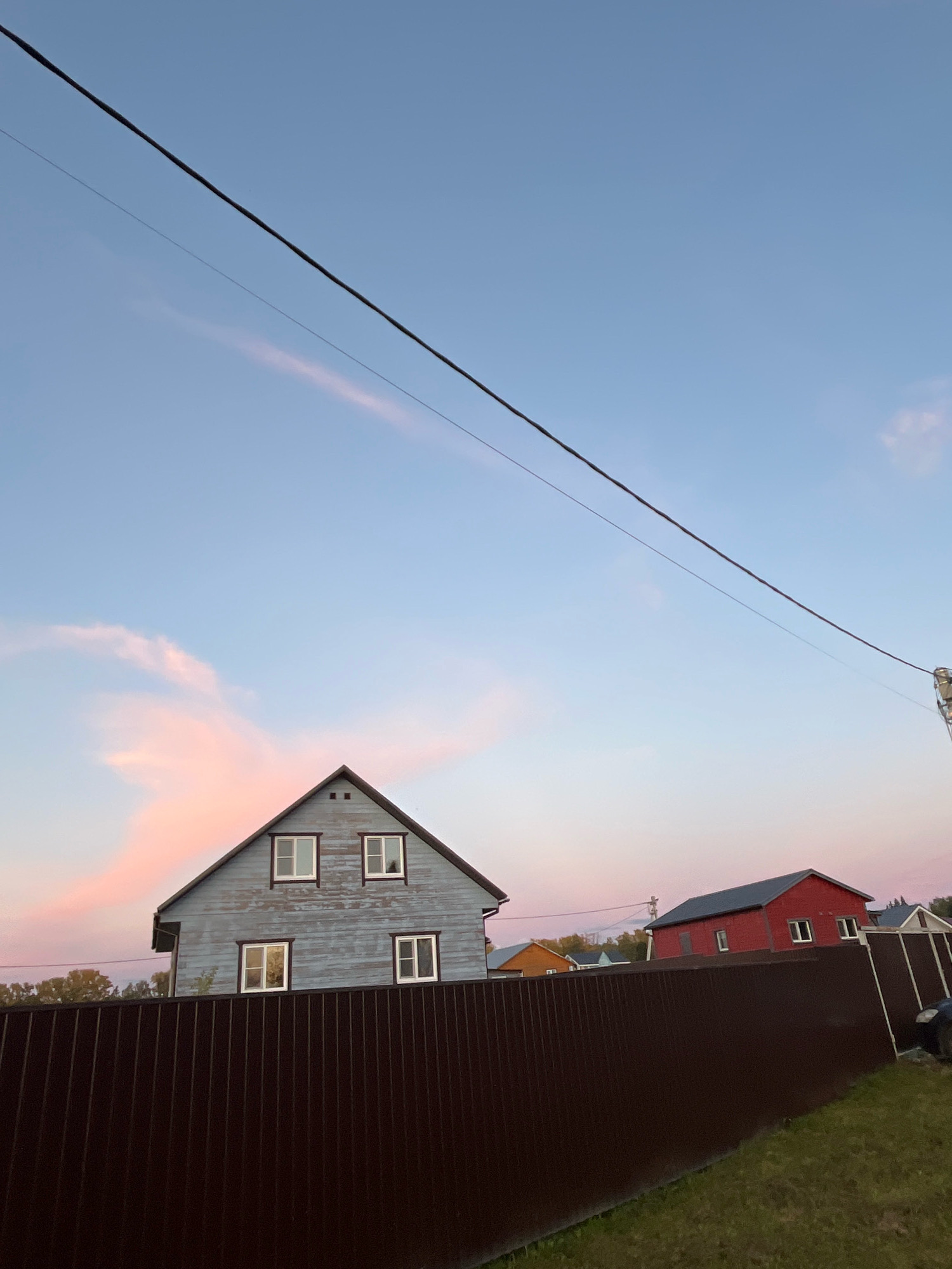 The image depicts a suburban or rural scene with several houses and a prominent fence in the foreground. The sky is a soft blue with streaks of pinkish-orange clouds, suggesting it is either sunrise or sunset. Two houses are clearly visible: a two-story house painted light blue with white-trimmed windows and a red two-story house. A dark brown, corrugated metal fence stretches across the lower half of the image, separating the viewer's vantage point from the houses. Green grass is visible on the right side of the fence. Overhead, a thick black power line stretches diagonally across the sky. The location is specified as Krasnaya Presnya SNT, Russia. There are no people visible in the image.