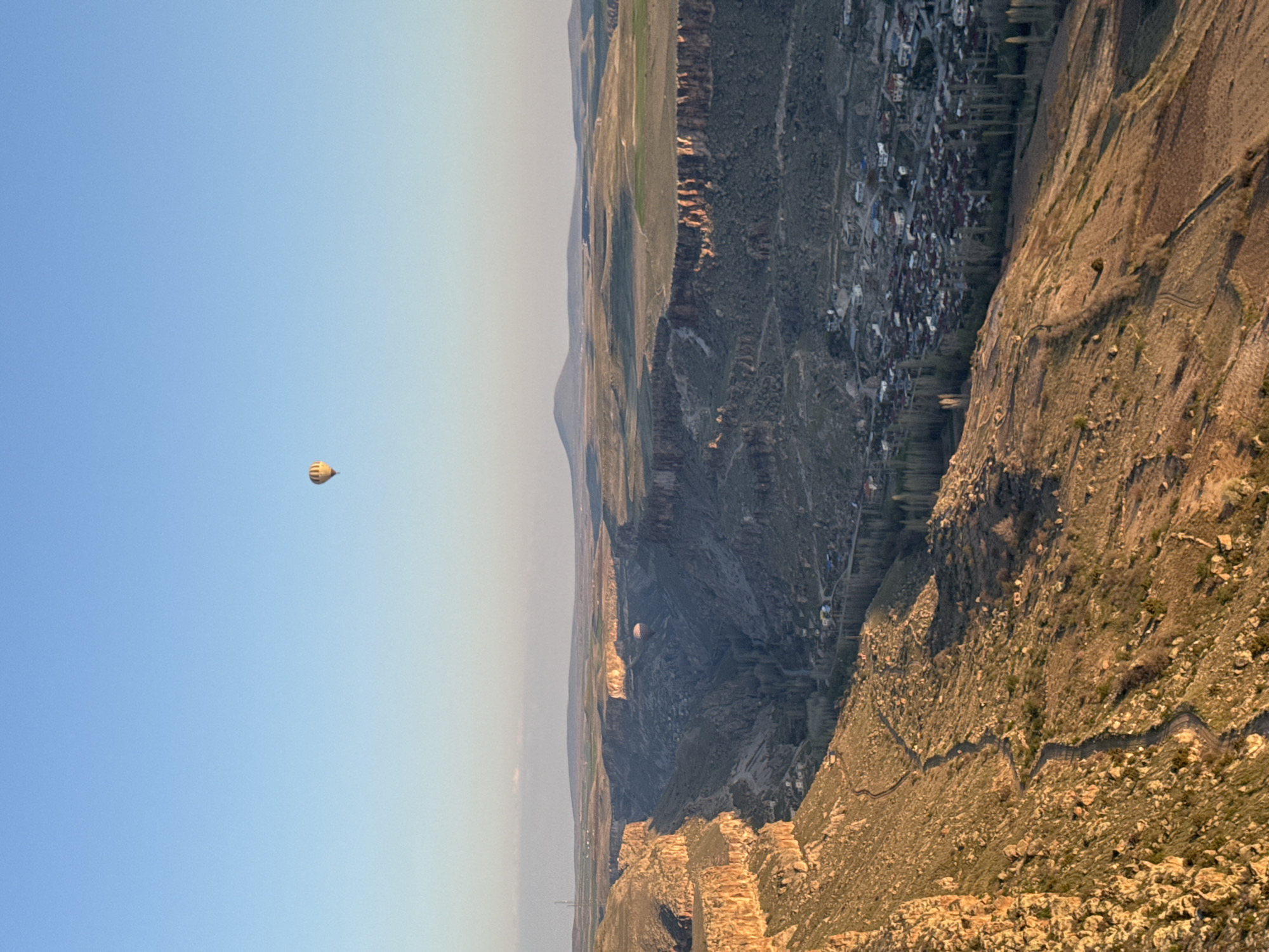 The image presents a panoramic aerial view of a rugged, expansive landscape, likely a deep canyon or valley, situated near Yeşilhisar, Turkey. The scene is dominated by dramatic geological formations under a clear, pale blue sky, suggesting either early morning or late afternoon due to the warm, golden light illuminating parts of the terrain.
In the upper-left portion of the sky, a distinct yellow hot air balloon with vertical stripes floats serenely. Another, smaller hot air balloon, lighter in color, is visible much further away, seemingly descending into or floating within the vast canyon.
The right side of the frame features a steep, rocky, and somewhat barren mountainside or cliff face, carved with winding paths or natural contours. This slope drops sharply into a deep valley. Across this valley, nestled against the opposite canyon wall, lies a small town or village characterized by numerous buildings and scattered trees, particularly noticeable around the settlement. Beyond the immediate canyon, the landscape extends into a series of undulating hills and distant mountains that fade into a hazy horizon. The overall impression is one of tranquility and grandeur, characteristic of a scenic hot air balloon flight over a majestic natural and historical landscape. No visible text is present in the image.