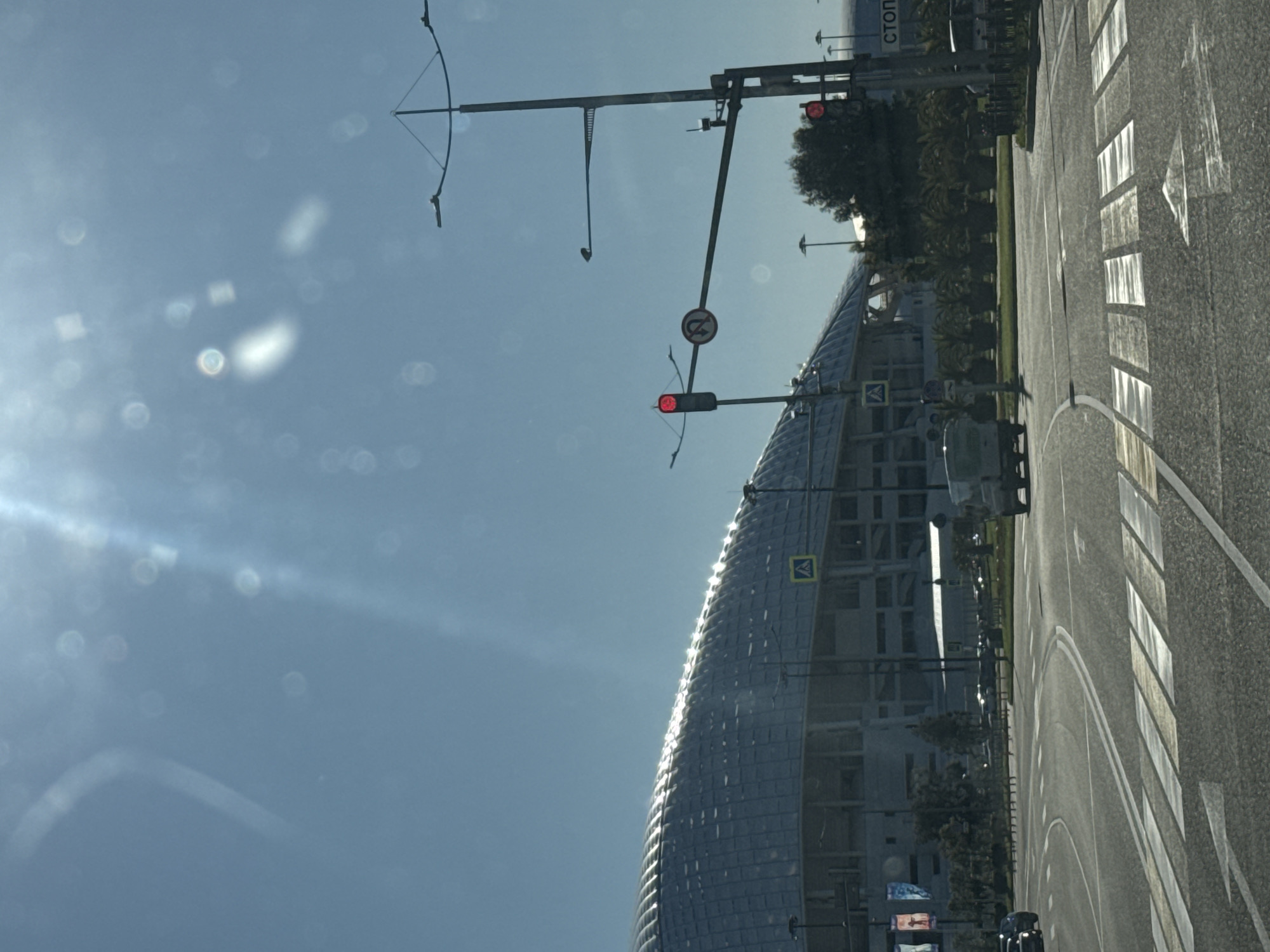 This image captures a bright, sunny daytime scene in Sirius, Russia, likely taken from a vehicle. The primary subject is a large, modern building with a distinctive curved, reflective facade, possibly a stadium or a large public venue, dominating the left side of the frame.
In the foreground, an asphalt road features clear white markings, including a pedestrian crossing and directional arrows. Overhead, a system of traffic lights and gantries spans the road, with a red traffic light illuminated, indicating a stop. A "No U-turn" sign (red circle with an arrow and diagonal line) is visible on one of the poles, alongside a yellow triangular "Pedestrian Crossing" sign. Partially visible in the background, on another pole, is a sign with the Russian text "СТОП" (STOP).
The sky is a clear blue, and strong sunlight creates prominent lens flares and reflections, suggesting the photo was taken through glass, such as a car windshield. Green foliage, including trees and bushes, lines the road and the area around the building. No people are visible, and the scene depicts a moment of urban transit on a clear day.