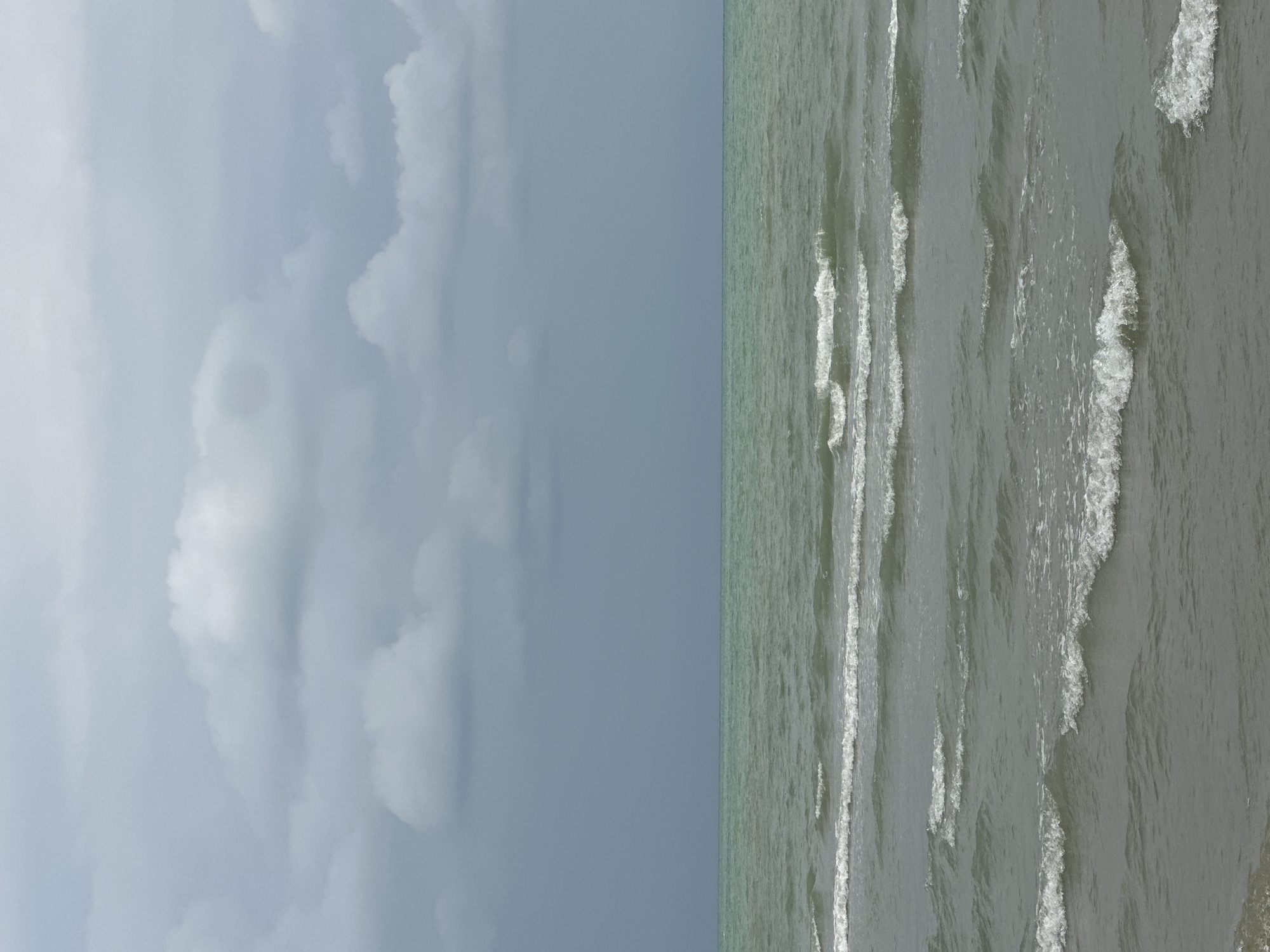 This is a vertical, wide-angle shot of a calm sea and a cloudy sky. The image is split diagonally, with the left half dominated by a soft grey sky filled with diffused clouds, suggesting an overcast day. The right half depicts the ocean with gentle, choppy waves that have white foam crests. The water appears a muted greenish-grey color, and the waves are moving towards the viewer from the right side of the frame. There are no people or man-made objects visible. The scene is tranquil and conveys a sense of peace. The lighting is diffused, indicating it is likely daytime, possibly late afternoon or early morning, given the soft quality of light. The overall impression is one of a serene coastal environment.