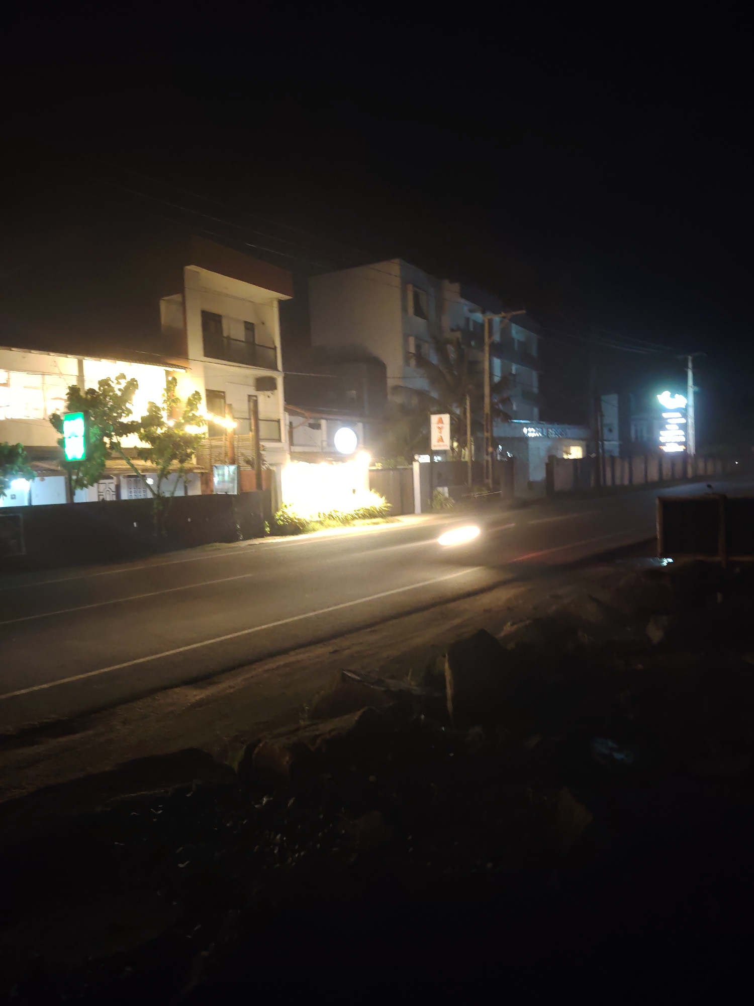 A nighttime, slightly blurry, wide shot shows a street in front of buildings. The street has a white line marking its lanes and a bright streak of light, indicating a moving vehicle's headlights, crossing from left to right. The buildings on the left are two stories with balconies and lit windows. A bright green rectangular sign is visible. Trees with illuminated leaves are planted in front of the buildings. Further down the street, a taller building with multiple floors and lit windows is present. A vertical sign with white lettering on a dark background stands on the right side of the street. The foreground is dark and features rough terrain with scattered rocks and debris. The sky is completely dark, suggesting it is late at night.