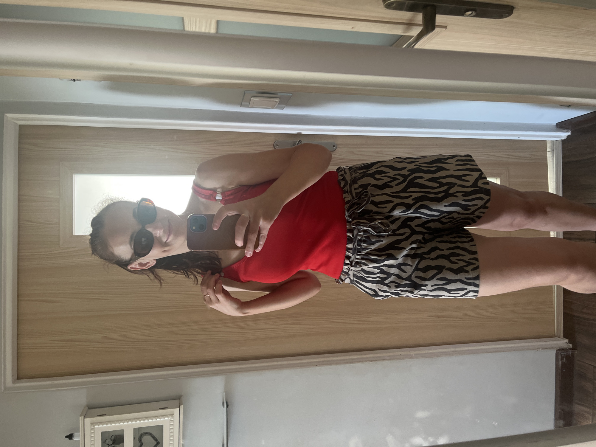 The image shows a young woman taking a mirror selfie. She is wearing a red tank top and black and white zebra-print shorts. She has sunglasses on and her hair is dark brown. She is holding a smartphone in her right hand, taking the picture. The background appears to be a hallway with a wooden door and doorframe. A light is visible through a window or glass panel in the door. The overall impression is casual and relaxed. The lighting suggests it is daytime.