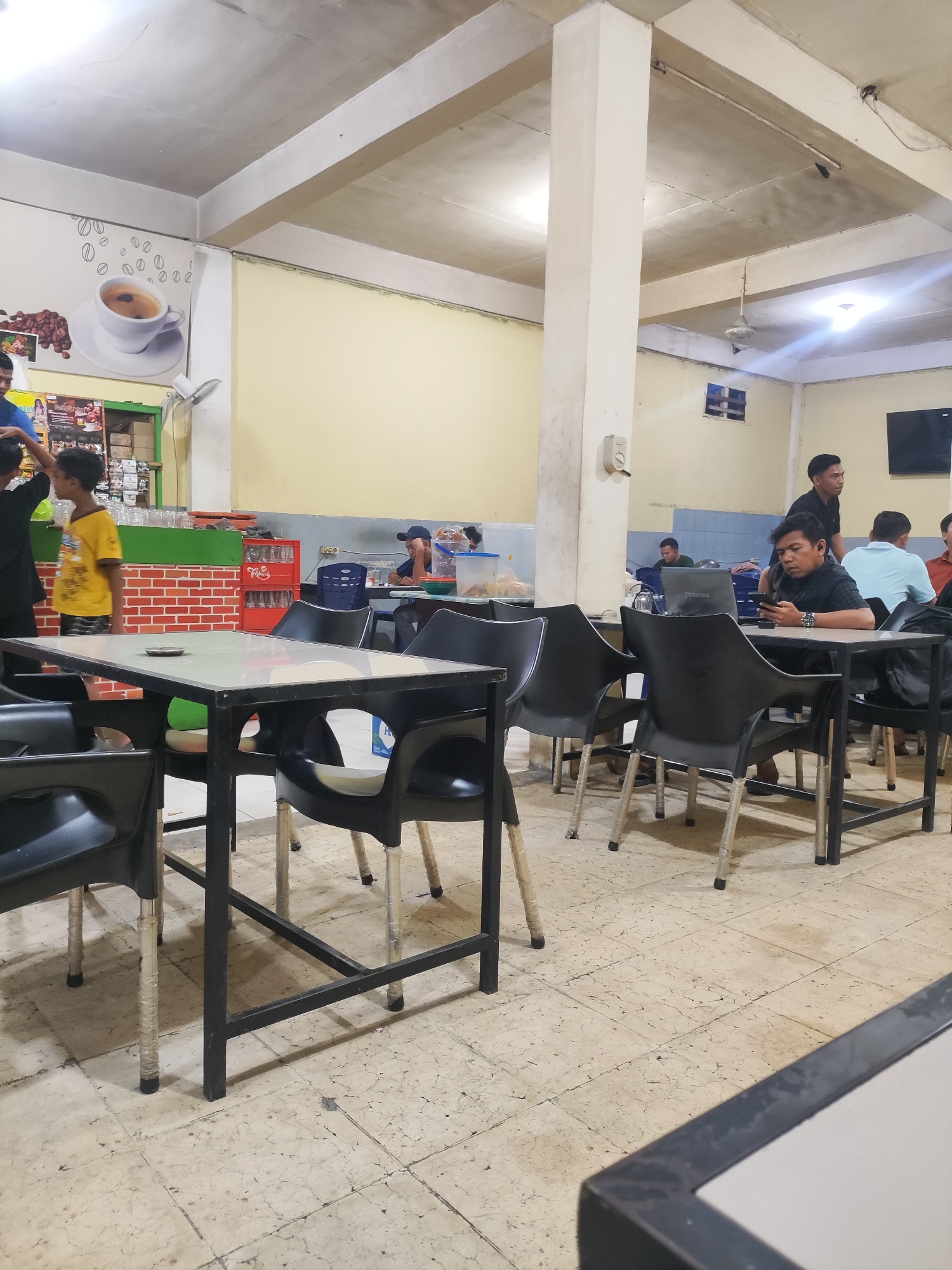 The image captures the interior of a casual eatery or cafe in Lhokseumawe, Indonesia, likely during the evening or night, indicated by the artificial lighting. The space features light yellow walls and a high ceiling with exposed concrete beams and several ceiling fans.
Numerous people, predominantly men and boys, are present. In the mid-ground, a man in a black plaid shirt is seated at a table, wearing earbuds and engrossed in his mobile phone, with an open "Toshiba" laptop in front of him. Behind him, another man in a black t-shirt stands near a wall-mounted television. Further back, several other individuals are seated at tables, some appearing to be socializing or engaged with their own devices. On the left side, near a green-topped counter with a red brick-patterned base, two boys and an adult are visible. One boy in a yellow shirt looks towards the camera, while another adult, partially obscured, wears a blue shirt. Stacked glasses and a red "Coca-Cola" crate are visible on or near the counter.
The establishment is furnished with practical black metal tables with light-colored tops and matching black plastic chairs; many chair legs are wrapped in clear plastic. A large mural on the left wall depicts coffee beans and a white coffee cup. The floor is covered with light-colored tiles, showing signs of wear and cracking. The overall atmosphere appears relaxed and informal, with patrons engaging in various leisurely activities like using electronics or conversing.