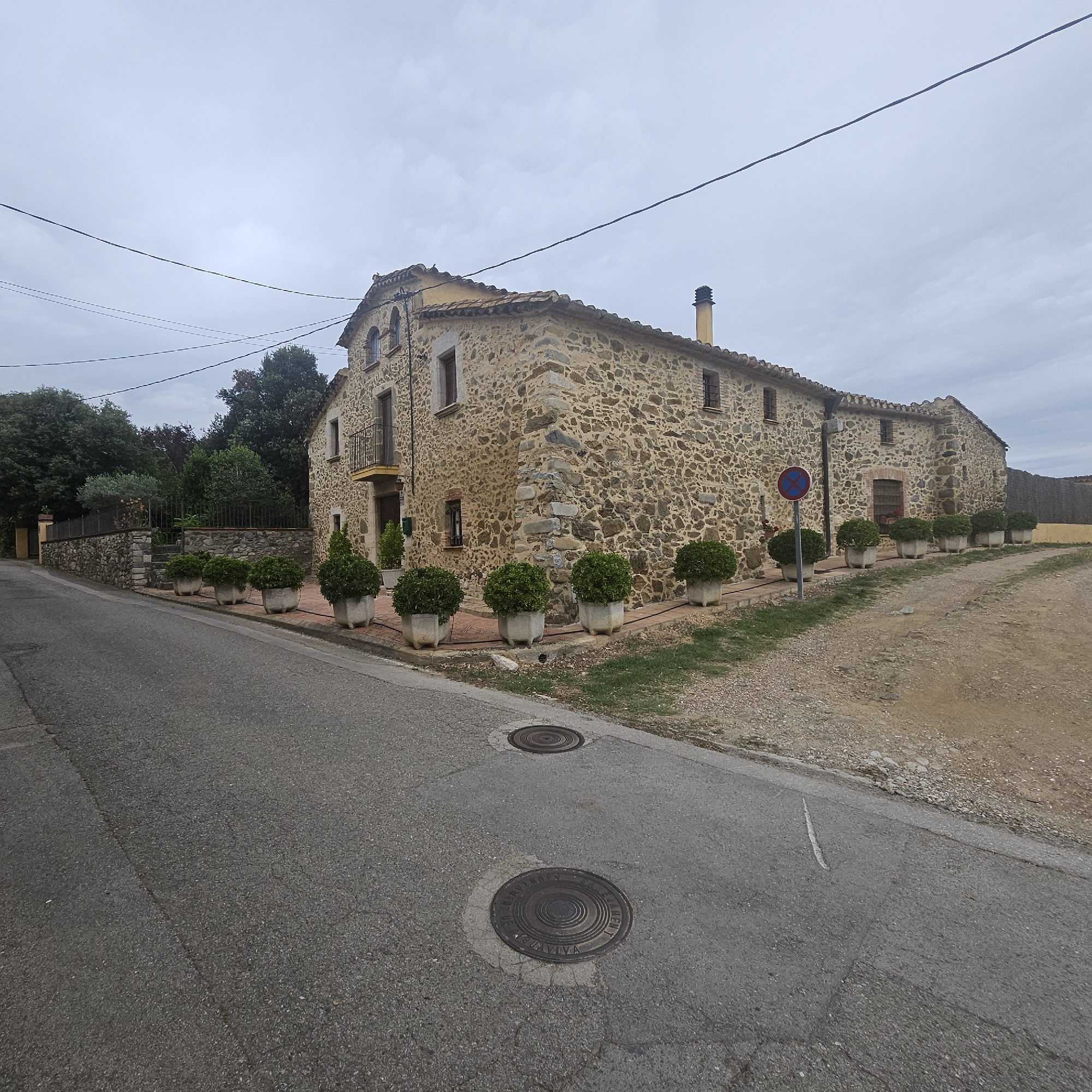 The image captures a rustic stone building in Mas Aliu, Spain, under an overcast sky. The building, likely a farmhouse, features a tiled roof, numerous small windows with dark frames, and a balcony with a wrought-iron railing on the second floor. A yellow accent color is visible around the balcony. A chimney stack protrudes from the roof. In front of the building, a line of potted green bushes adds a touch of manicured greenery along a brick pathway. To the left, a stone wall separates the property from a dense cluster of trees and foliage. A paved road curves in the foreground, with two circular manhole covers visible on its surface. A gravel path borders the road on the right, leading towards the side of the stone structure. A circular traffic sign with a blue background and red markings is positioned near the building. The overall scene conveys a rural, somewhat aged aesthetic. There are no people visible in the image.