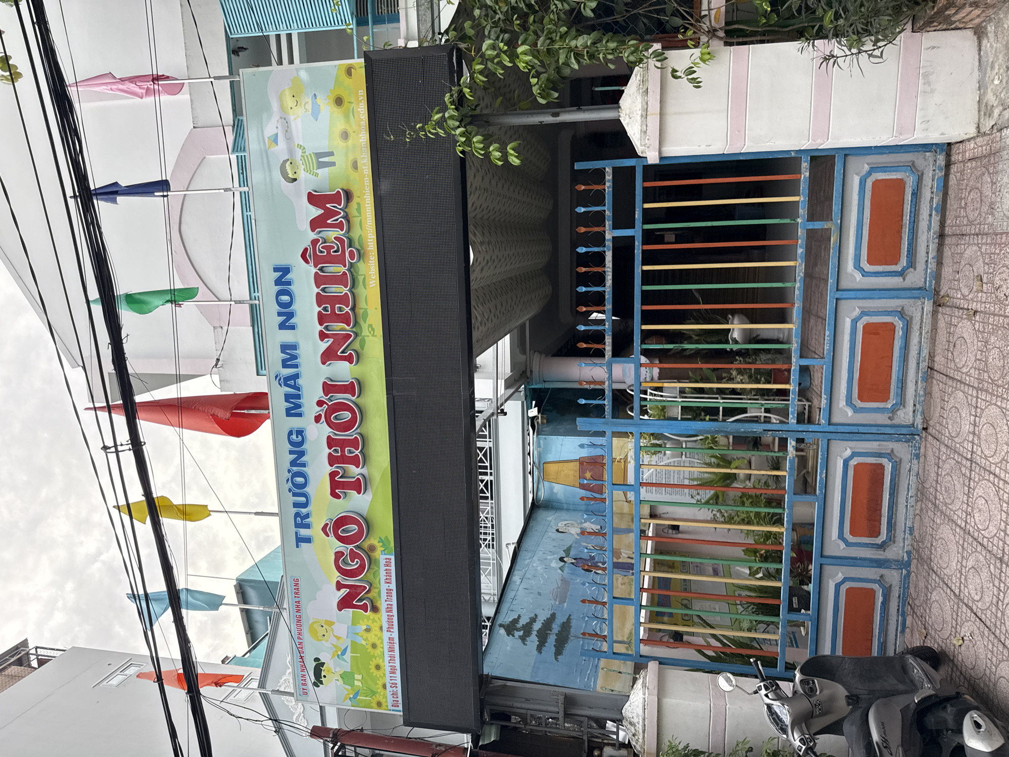 The image shows the exterior of a kindergarten in Nha Trang, Vietnam. A large sign in Vietnamese, reading "TRƯỜNG MẦM NON NGÔ THỜI NHIỆM" (Ngo Thoi Nhiem Kindergarten), is prominently displayed. Below the sign is a black LED screen, currently blank. The entrance features a colorful metal gate with vertical stripes of blue, red, yellow, and green, and a mural on the wall behind it depicting a seascape with ships and people. A scooter is parked to the left of the entrance, and a white pillar with pink stripes stands to the right. Five flags of various colors (orange, blue, yellow, green, dark blue, and pink) are visible on poles atop the building, against a cloudy sky. The kindergarten appears to be located on a street in a densely populated urban area, suggested by the adjacent buildings and overhead electrical wires.