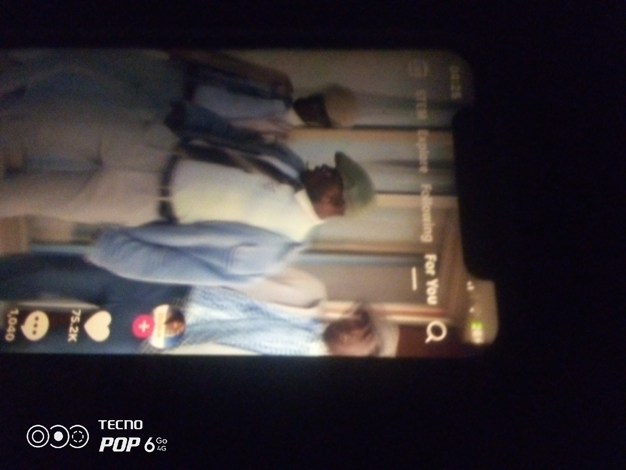 The image is a low-resolution, vertically oriented photograph of a smartphone screen displaying a social media feed, likely TikTok, given the icons present. The content on the screen appears to be a video featuring at least two individuals. The primary subject in focus is a person wearing a light-colored outfit and a light green cap, looking down. Behind them, another person is visible, also in light-colored clothing. The environment within the video appears to be indoors. On the right side of the screen, vertical text is partially visible, indicating categories like "Following," "Explore," and "For You," characteristic of social media platforms. At the bottom of the screen, there are icons for likes, comments, and shares, with numerical indicators. The phone itself has the "TECNO POP 6 4G" branding at the bottom left, suggesting the device it is being viewed on. The image is captured in a dark environment, indicating the photo was likely taken at night or in a dimly lit room, with the smartphone screen being the primary light source. There is no discernible information in the image that would indicate a specific location in the city of Howayi, Nigeria.