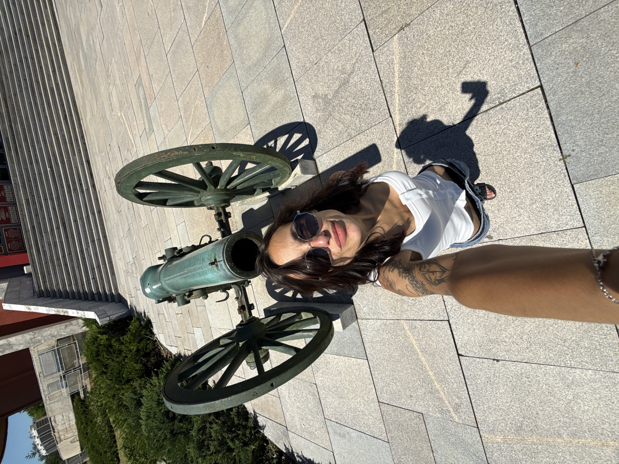 The image shows a woman taking a selfie in front of an old cannon. She is wearing a white top, blue jeans, and sunglasses. The cannon is green and mounted on a carriage with two large wheels. Behind the cannon, there is a wide staircase leading up to a building. The ground is paved with large stone tiles. The location is the Pleven Panorama, a monument complex in Pleven, Bulgaria, commemorating the Siege of Plevna during the Russo-Turkish War.