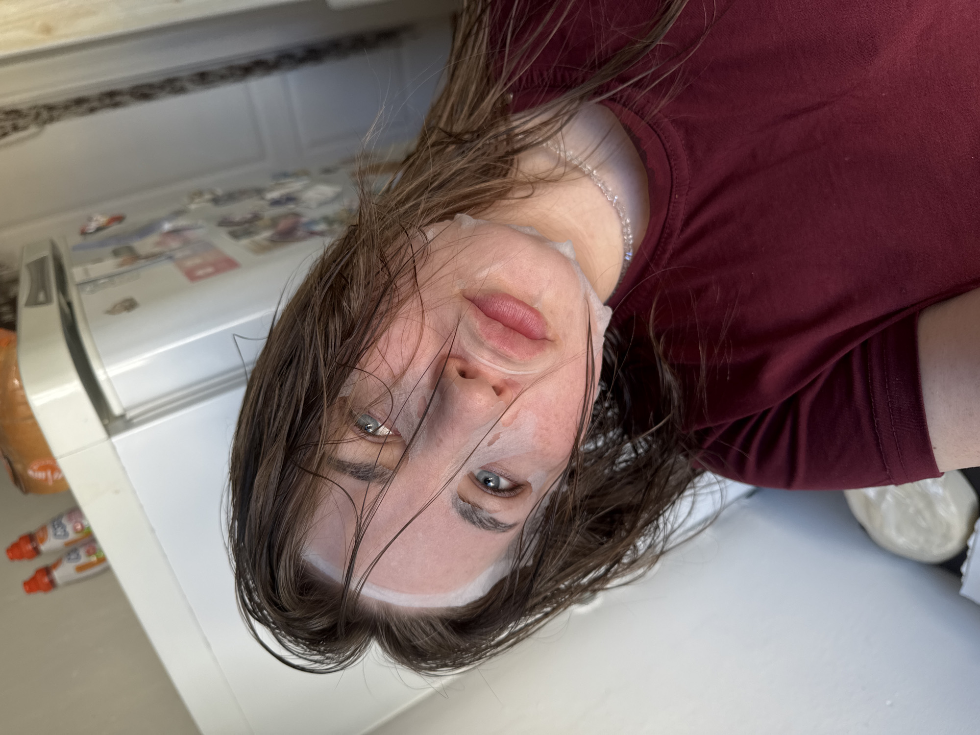 A woman is wearing a sheet mask and a maroon shirt while in a kitchen. The kitchen has a refrigerator and a countertop. There is a loaf of bread on the countertop. The location is assumed to be Chaikovsky, Russia.