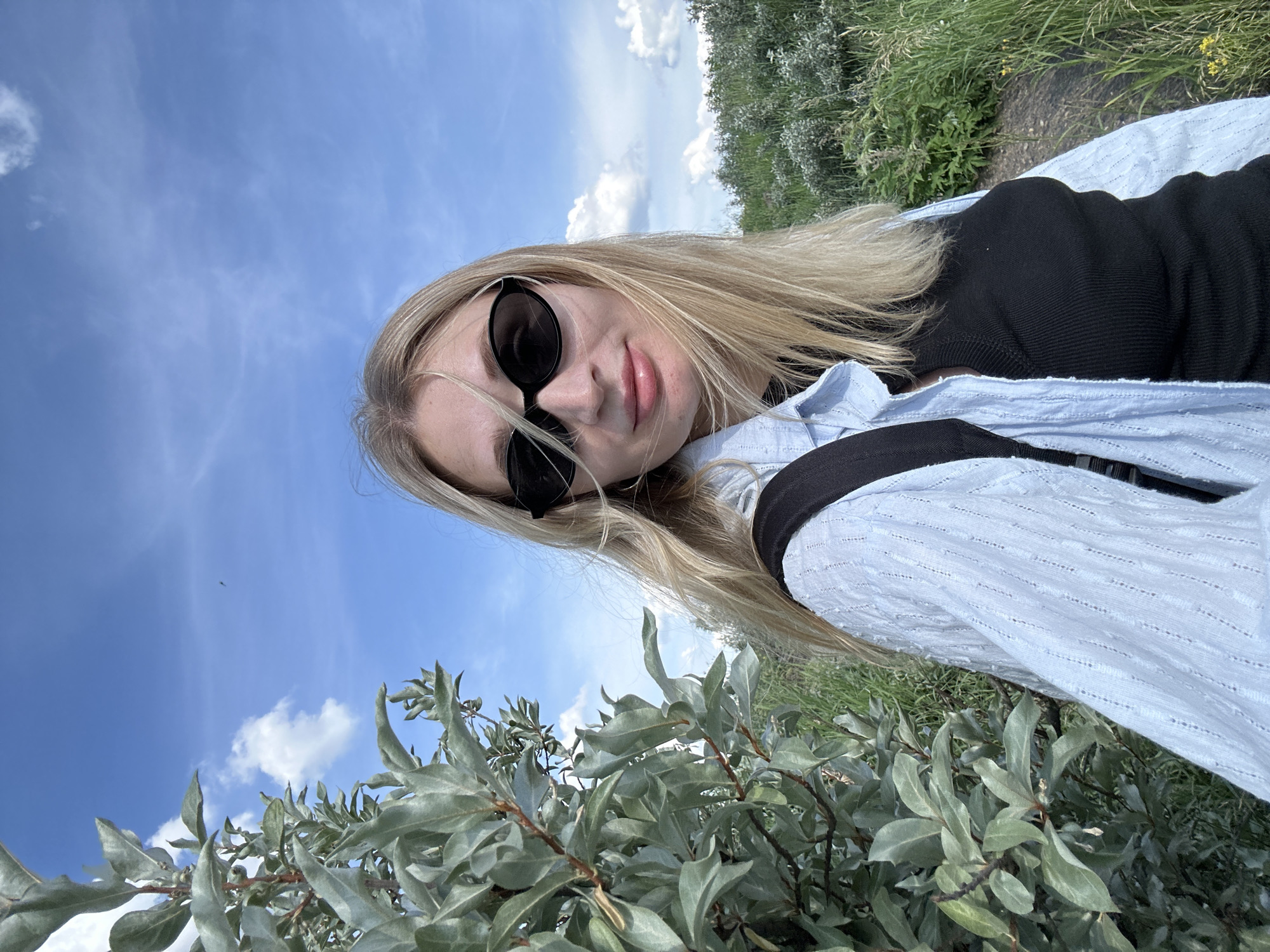 A young woman with blonde hair and sunglasses is taking a selfie outdoors on a cloudy day. She is wearing a light blue button-down shirt over a black top and a backpack. To her left is a shrub with silvery-green leaves. In the background, there is tall green grass and a path. The sky is blue with scattered white clouds. This image was taken near the Kama River in Chaikovsky, Russia.