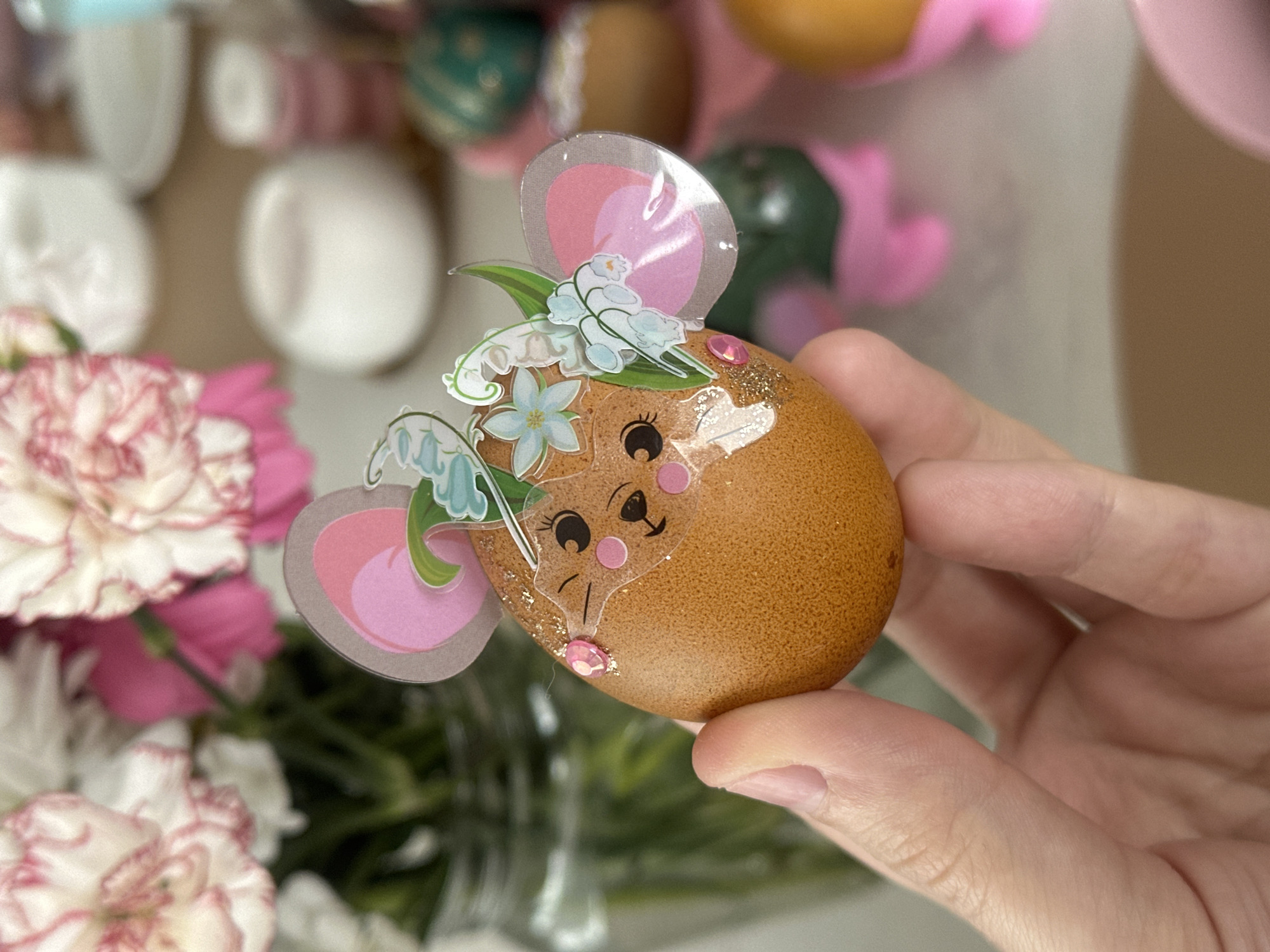 A human hand is holding a brown egg decorated to resemble a mouse. The egg has stickers affixed, including ears outlined in grey with pink centers, a mouse face with black eyes and a small triangular nose, and floral decorations featuring white flowers and green leaves. A small pink gemstone is affixed to the tip of the egg. Gold glitter is sprinkled near the base of the mouse face. The background is blurry, but shows various objects including pink, green, and brown Easter eggs, a vase with white and pink carnations, and white decorative objects. The location is not determinable from the image.
