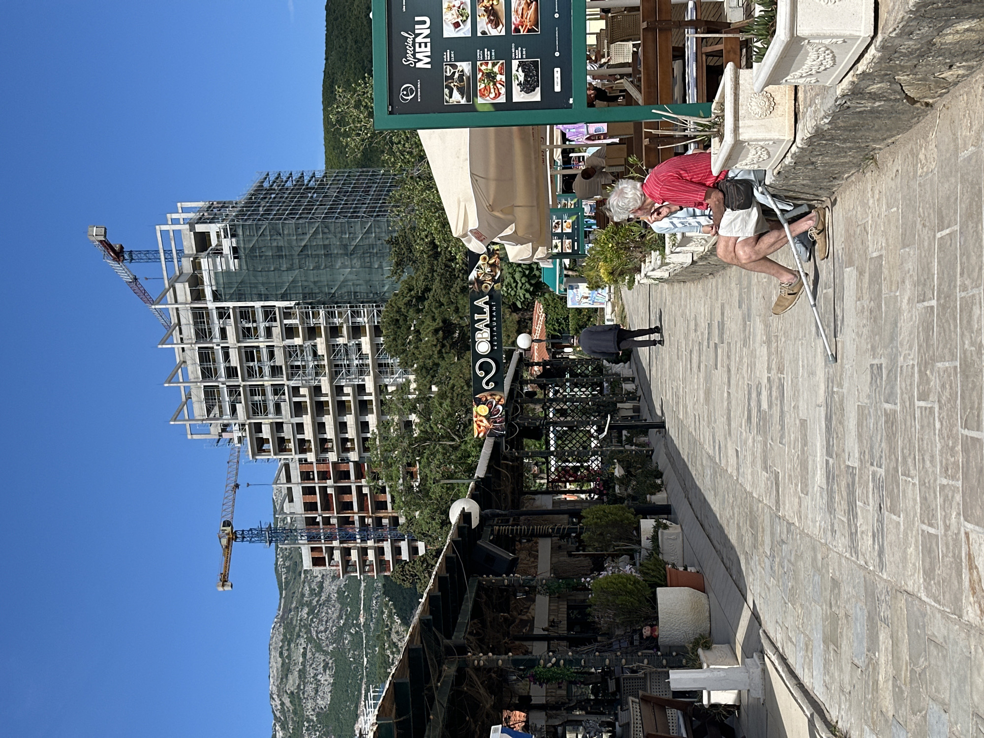 The image shows a street scene in Budva, Montenegro, with a building under construction in the background. A man with gray hair and a red shirt is leaning against a wall, using a crutch. Behind him is a restaurant sign that reads "OBALA RESTAURANT" and a "Special Menu" board. Several people are seated at tables in the restaurant area. In the background, a large, unfinished building with scaffolding and two cranes dominates the skyline. The sky is clear and blue.