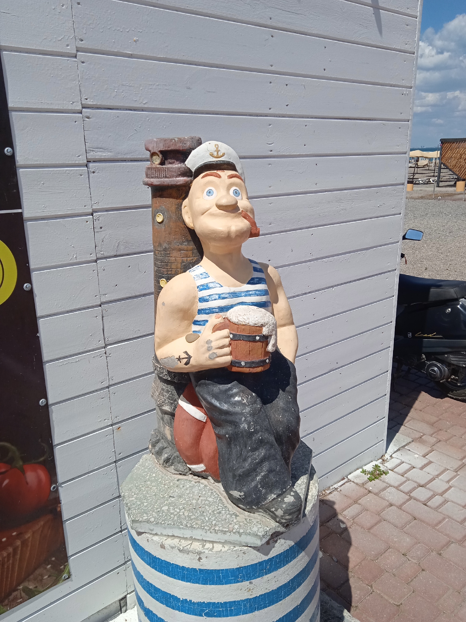 In Novofedorovka, Ukraine, a statue of a cartoonish sailor wearing a striped shirt, white cap with an anchor, and holding a wooden mug of foamy beer is positioned next to a light grey wooden building. The sailor has rosy cheeks, a pronounced nose, and a pipe in his mouth. He has a tattoo of an anchor on his left forearm. The statue sits on a cylindrical base painted with blue and white stripes, which is mounted on a concrete pedestal. To the right of the statue, a black scooter is parked on a brick pathway. In the background, a beach area with lounge chairs and a wooden structure is visible under a partly cloudy sky.