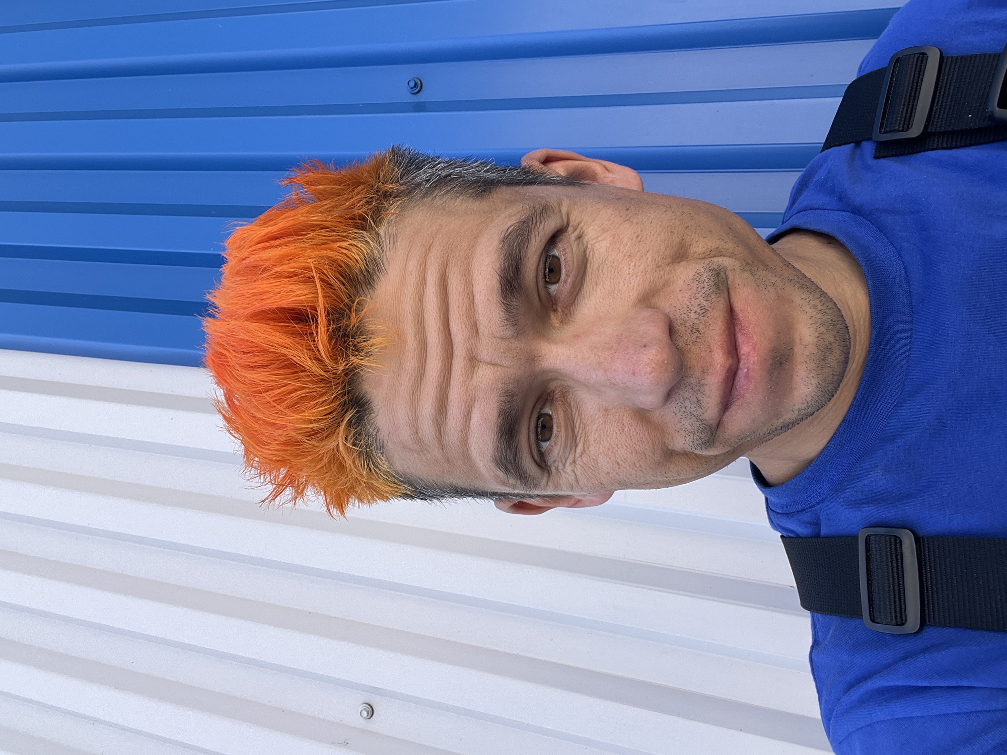 The image shows a close-up selfie of a man with spiky, bright orange hair. He is wearing a blue t-shirt and black suspenders. The background is a corrugated metal wall divided vertically into white and blue sections. The man's expression is a slight smile. The location is Chaikovsky, Russia.
