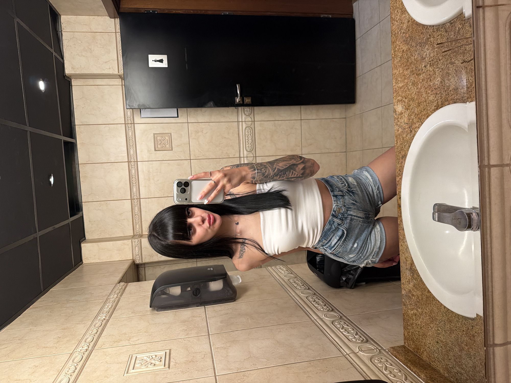A young adult female is depicted taking a mirror selfie in a public restroom located in the city of Plovdiv, Bulgaria. She has long, straight black hair with bangs and is wearing a white strapless crop top and light blue distressed denim shorts. Her right arm (visible as her left in the mirror) is extensively tattooed with a full sleeve design, and a smaller tattoo is visible on her left shoulder/chest area (visible as her right in the mirror). She holds a silver smartphone in her right hand (visible as her left in the mirror) to capture the image, displaying multiple rings on her fingers and a silver bracelet on her wrist. Her expression is a slight pout.
The restroom features light beige tiled walls with a decorative horizontal border and occasional framed square tiles. The ceiling is a dark grey/black false ceiling with visible grid lines and recessed lighting fixtures. A dark grey/black paper towel or soap dispenser is mounted on the left wall, with a black waste bin partially visible beneath it. On the right, a black door with a silver handle displays a white female restroom symbol. The foreground shows a speckled brown and tan granite or laminate countertop with two partially visible white ceramic sinks and chrome faucets. The floor is covered in lighter beige tiles.
