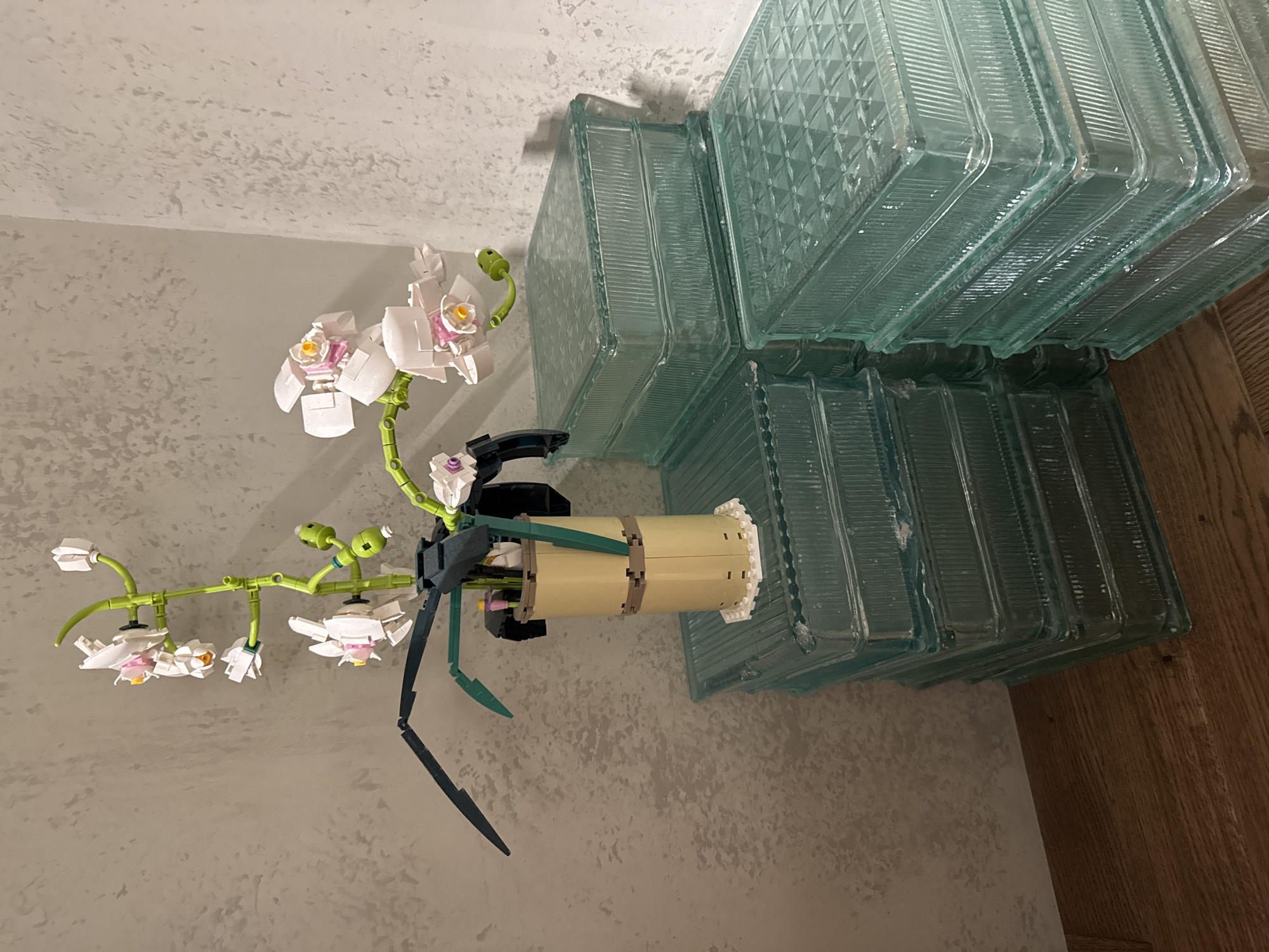 A close-up, eye-level shot focuses on a Lego orchid plant arranged in a beige pot, positioned on top of stacks of clear, square glass blocks. The orchid, constructed from Lego bricks, features white and pink flowers with yellow centers, green stems, and dark green leaves. The pot itself is a cylindrical Lego build with a white circular base. The glass blocks are stacked in two distinct piles, one slightly higher than the other. The background consists of a textured, light-colored wall meeting at a corner. The floor is made of wooden planks. The scene is likely an indoor setting.