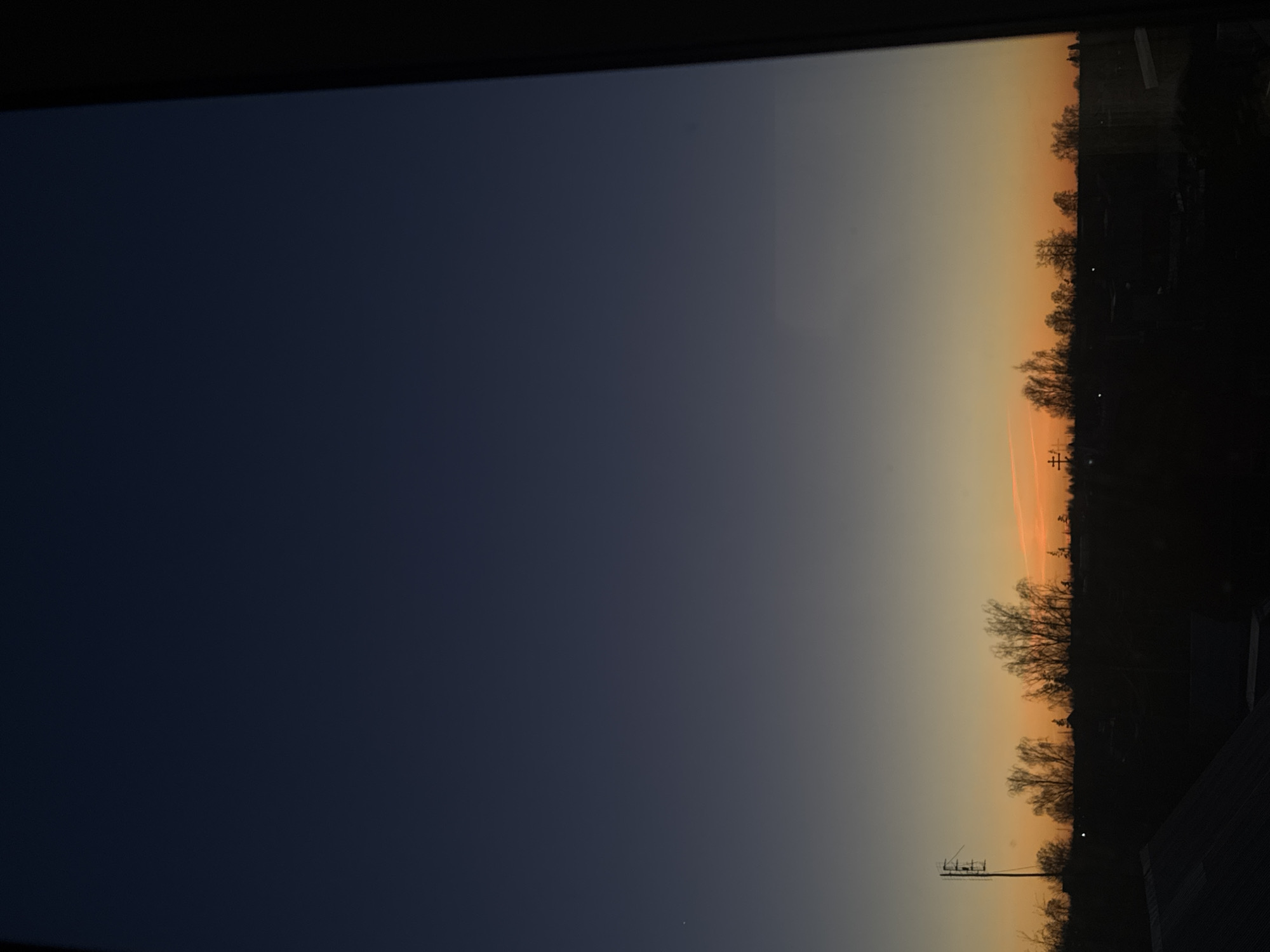 Captured in Фоки, Russia, the photograph depicts a landscape transitioning from dusk to night. The left portion of the frame displays the deepening blue of the evening sky, contrasted against the right side where the sunset lingers, painting the sky with shades of orange and yellow. Silhouetted against the fading light, a line of trees and distant buildings creates a distinct horizon. Lower in the frame, an antenna stands out, a clear marker of human presence amidst the natural scene.