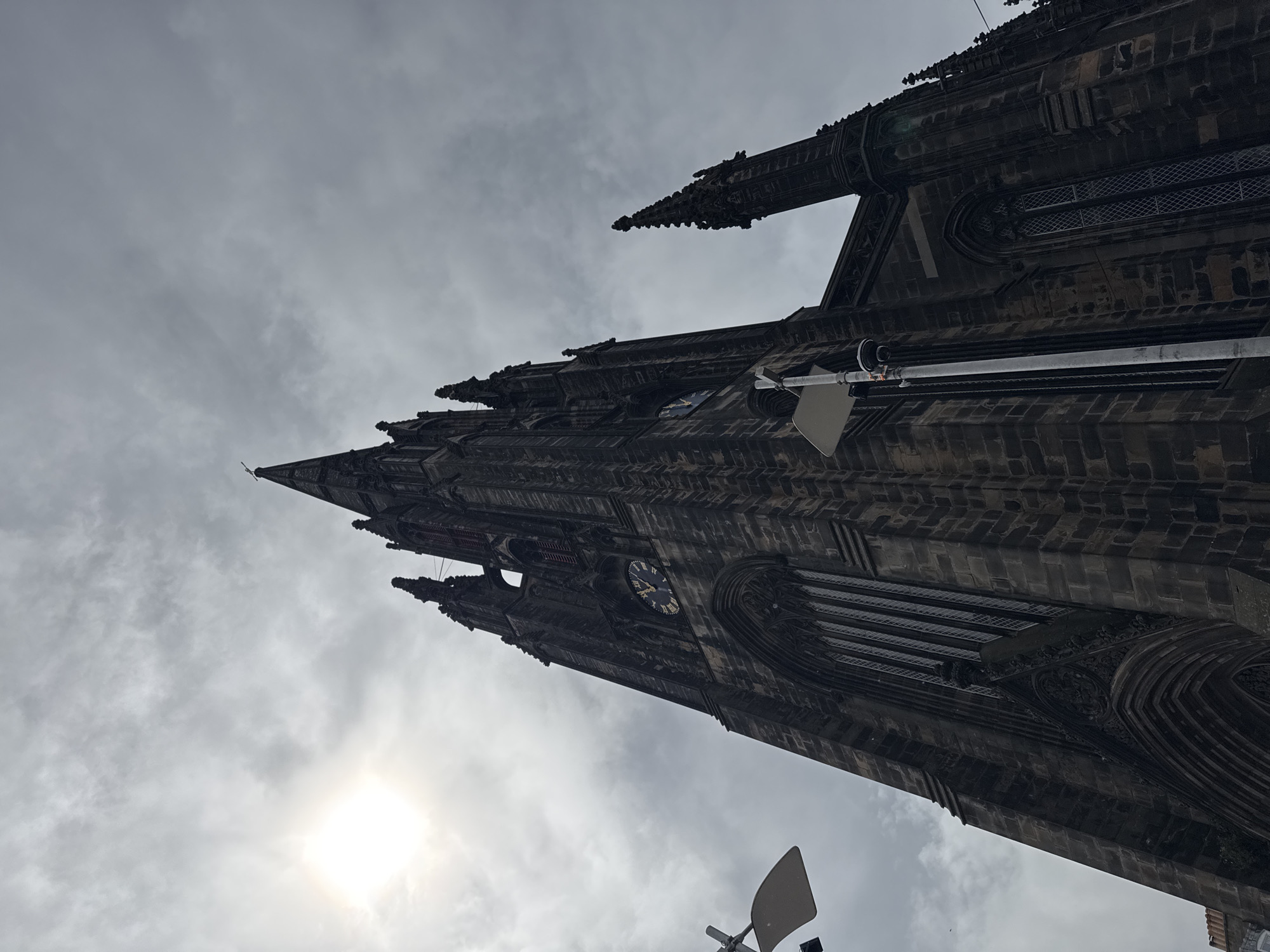 A low-angle shot captures the imposing facade of Tolbooth Kirk, a historic church located in the City of Edinburgh, United Kingdom. The dark, Gothic architecture of the church dominates the frame, with its spires reaching towards a cloudy, overcast sky. The sun peeks through the clouds on the left side of the image, casting a soft glow. A large clock face is visible on the church tower, its roman numerals suggesting the time. Streetlights and a security camera are positioned in the foreground, partially obscuring the church's details. The overall mood is one of solemn grandeur, characteristic of Edinburgh's historic cityscape.