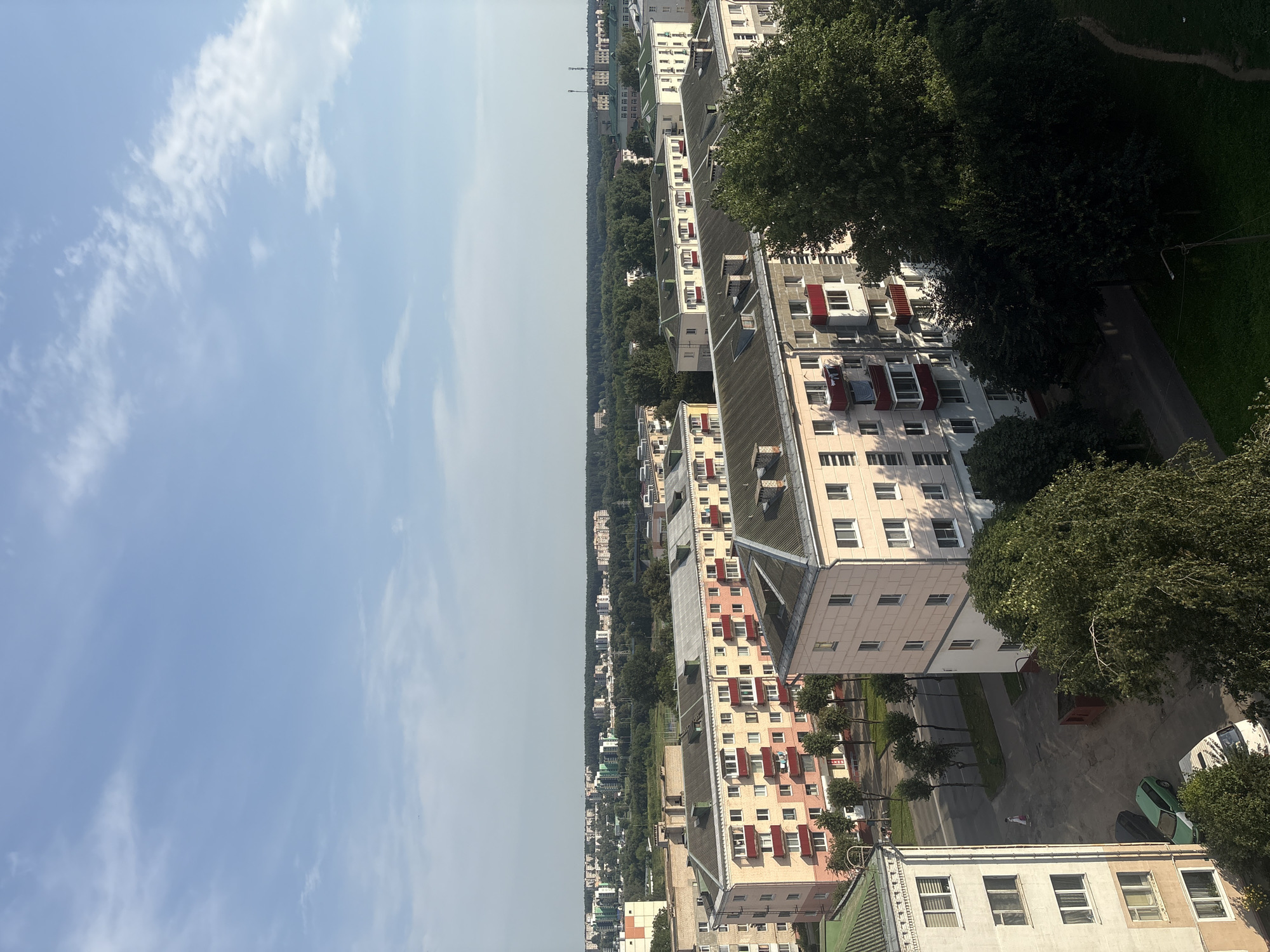 The image is an aerial view of a residential area in Orsha, Belarus, on a clear day with a few wispy clouds. In the foreground, there are several multi-story apartment buildings, characterized by their light-colored facades and red balconies. The buildings appear to be of Soviet-era architecture. A street runs between some of the buildings, with parked cars, including a green car and a white van. A single person in a light-colored outfit is walking on the sidewalk. Dense green trees are prominent throughout the scene, especially to the right of the image, and line the streets. In the background, more apartment buildings and industrial structures can be seen interspersed with a thick band of forest. The overall impression is of a typical urban residential district in Eastern Europe.