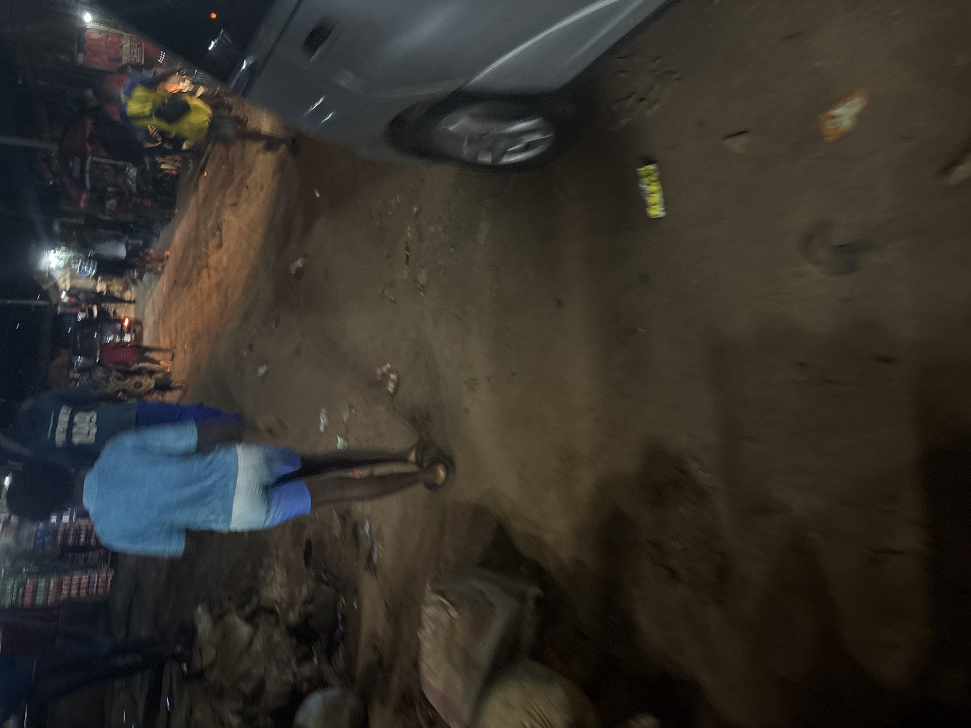 The image depicts a street scene at night in Nkpor, Nigeria. A young person wearing a blue t-shirt and white shorts is walking away from the camera. In the background, several other individuals are present, some appearing to be shopkeepers or patrons in what looks like a market area. There are stalls or shops with items displayed, and a silver vehicle is parked on the right side of the frame. The ground is dirt and littered with debris. The lighting suggests it is dimly lit, likely from streetlights or shop illumination.