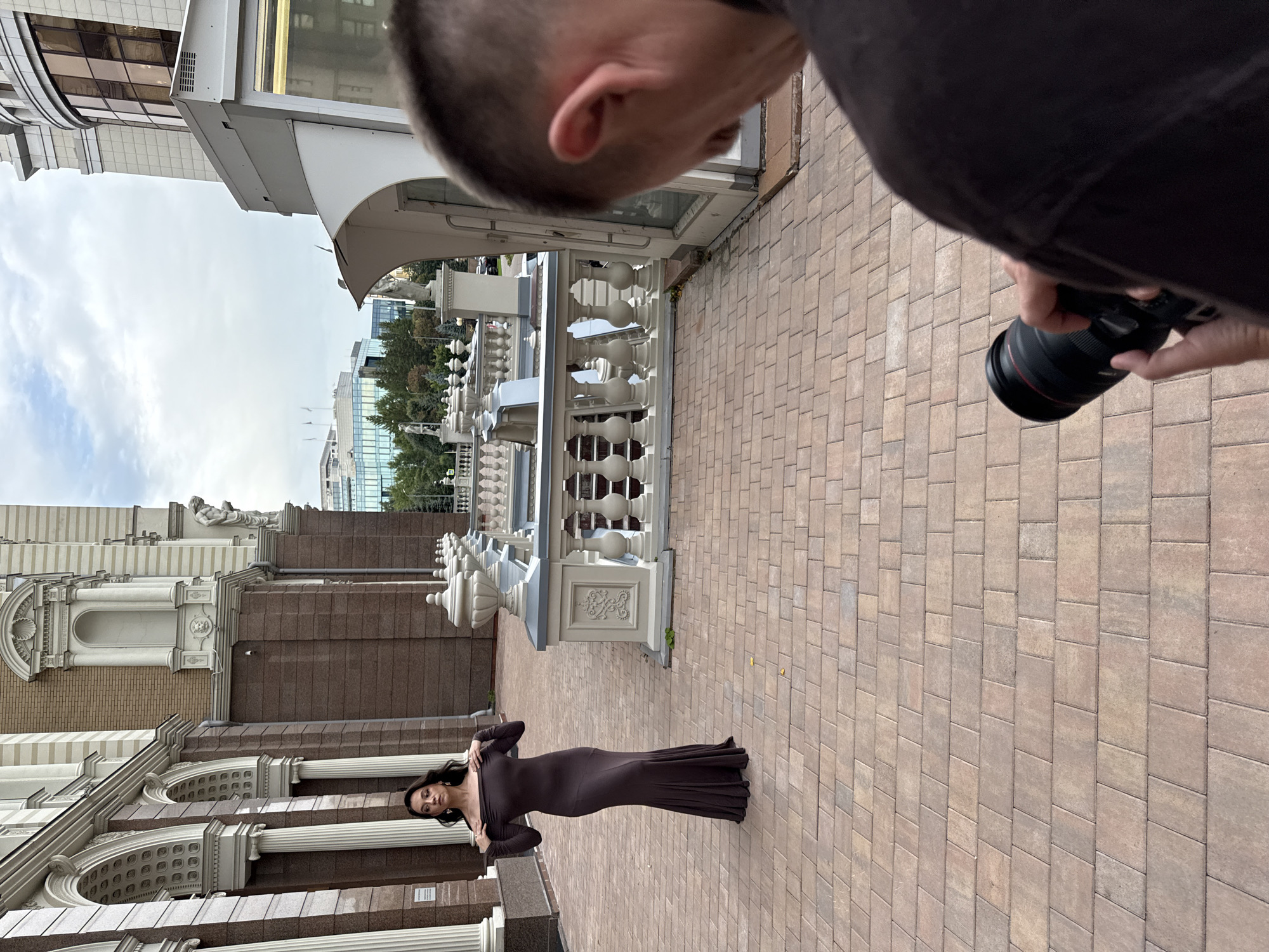 A woman in a long, dark brown, off-the-shoulder dress stands on a brick-paved walkway in front of an ornate, classical-style building with columns and arched windows. She is looking towards the camera with her hands near her neck. In the foreground, the back of a person's head and shoulders are visible, holding a camera with a large lens. The background shows a modern glass-fronted building and a cloudy sky. The overall scene appears to be an outdoor photoshoot.