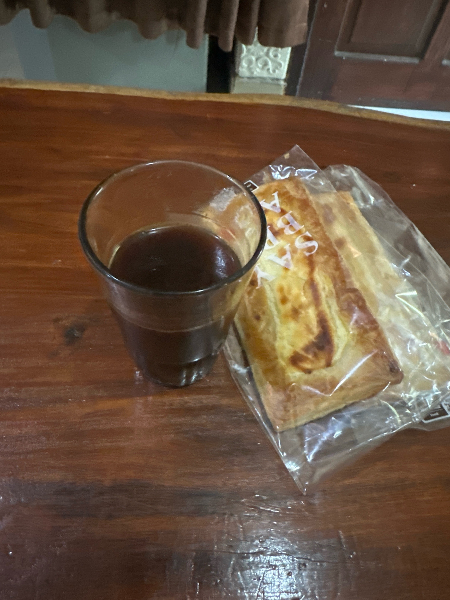 A glass of dark liquid, likely coffee, sits on a wooden table next to a cellophane-wrapped pastry. The pastry appears golden brown and has a white, curved design on its surface. The table's surface has a visible wood grain and reflects light. In the background, a blurred element suggests a curtain or drapery above a light-colored wall, possibly indicating an indoor setting. The overall impression is a simple still life, suggestive of a snack or breakfast. The image does not contain any specific landmarks or features that would allow for its precise localization within Selemadeg Barat, Indonesia, but it depicts a common domestic or cafe setting.