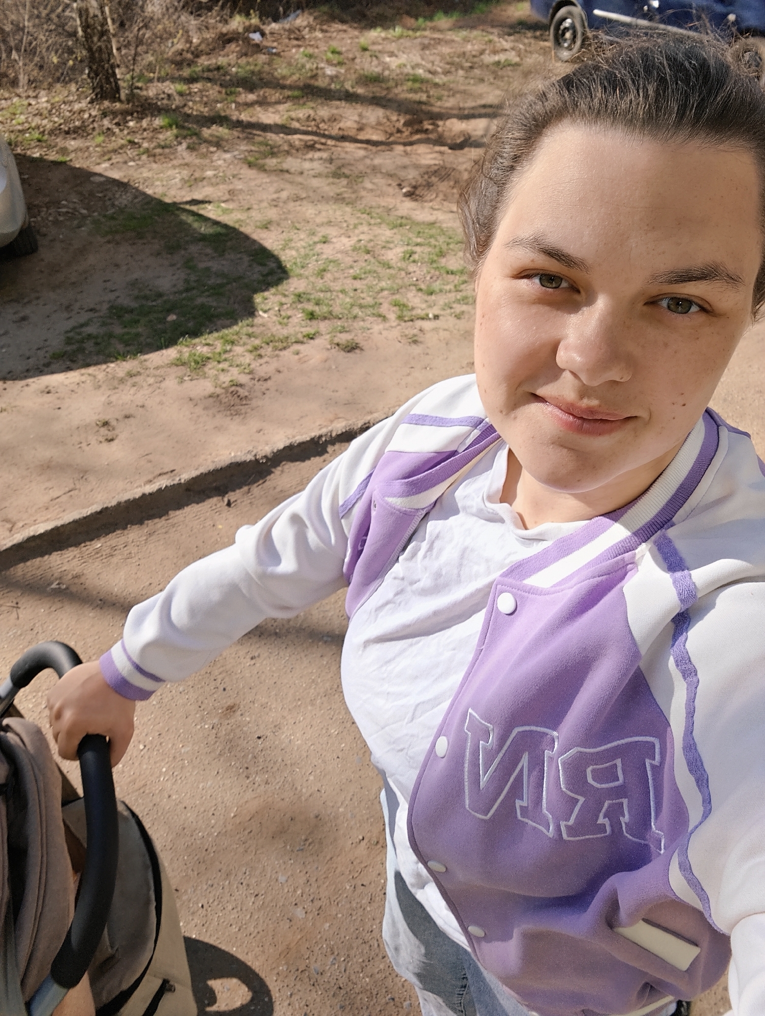 A light-skinned woman with her hair pulled back stands outdoors, likely holding a stroller, as indicated by the black handle in the lower left corner. She is wearing a white t-shirt and a white and lavender letterman-style jacket with the letters "NR" visible. The ground appears to be a dirt or gravel path. Patches of grass are visible near the edge of the path and near a tree in the background. Part of a vehicle is visible in the upper right corner, and a car is parked in the upper left corner. The photograph was taken during the day in a location in the city of Chaikovsky, Russia.