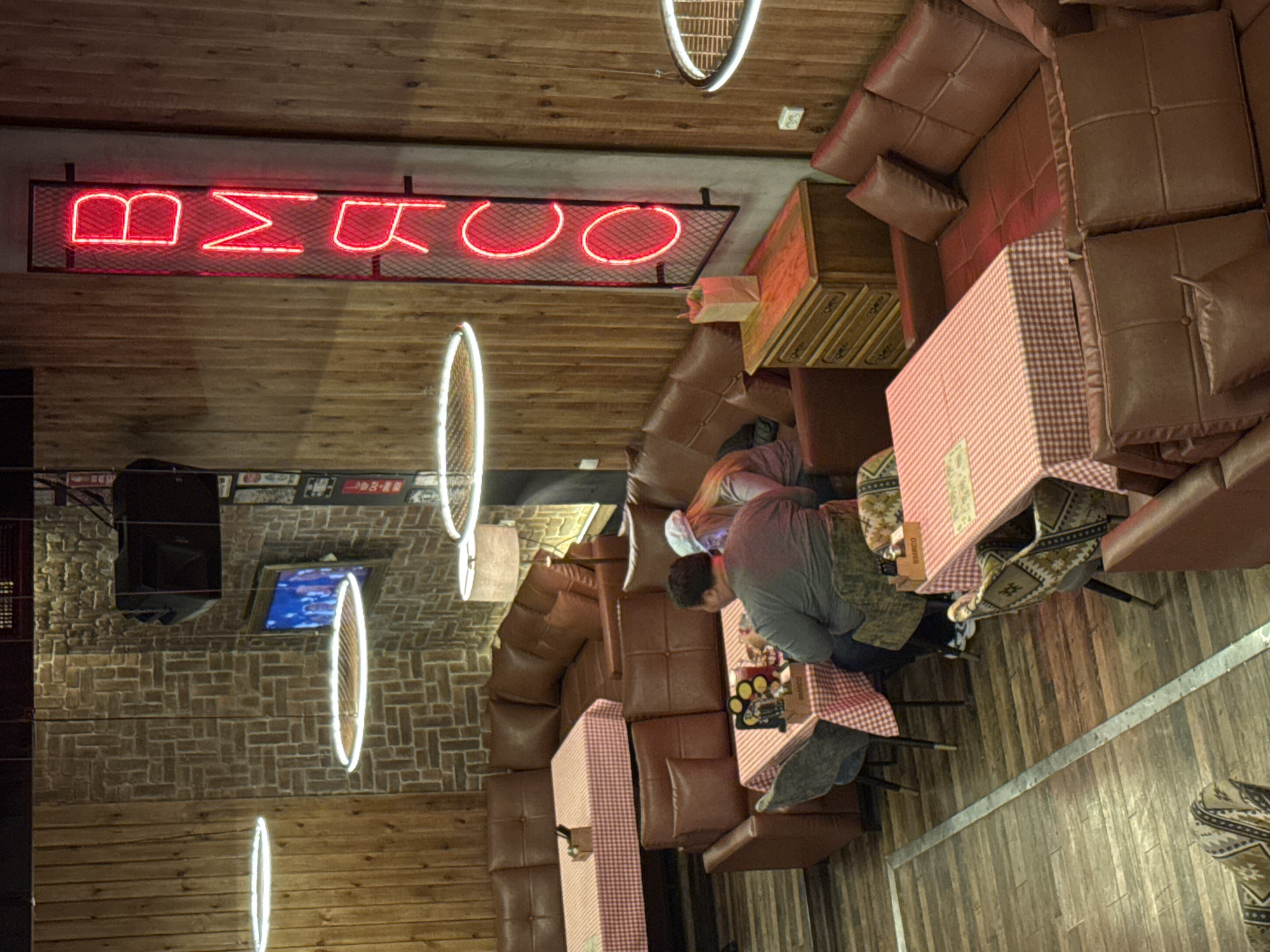 Here is a description of the image.
The image depicts the interior of a restaurant or cafe. A red neon sign with the text "BMRCO" is displayed on a wooden-paneled wall. The cafe is equipped with brown leather seating booths with checkered tablecloths. Patrons are seated at the tables. At one table, two people are having a meal.