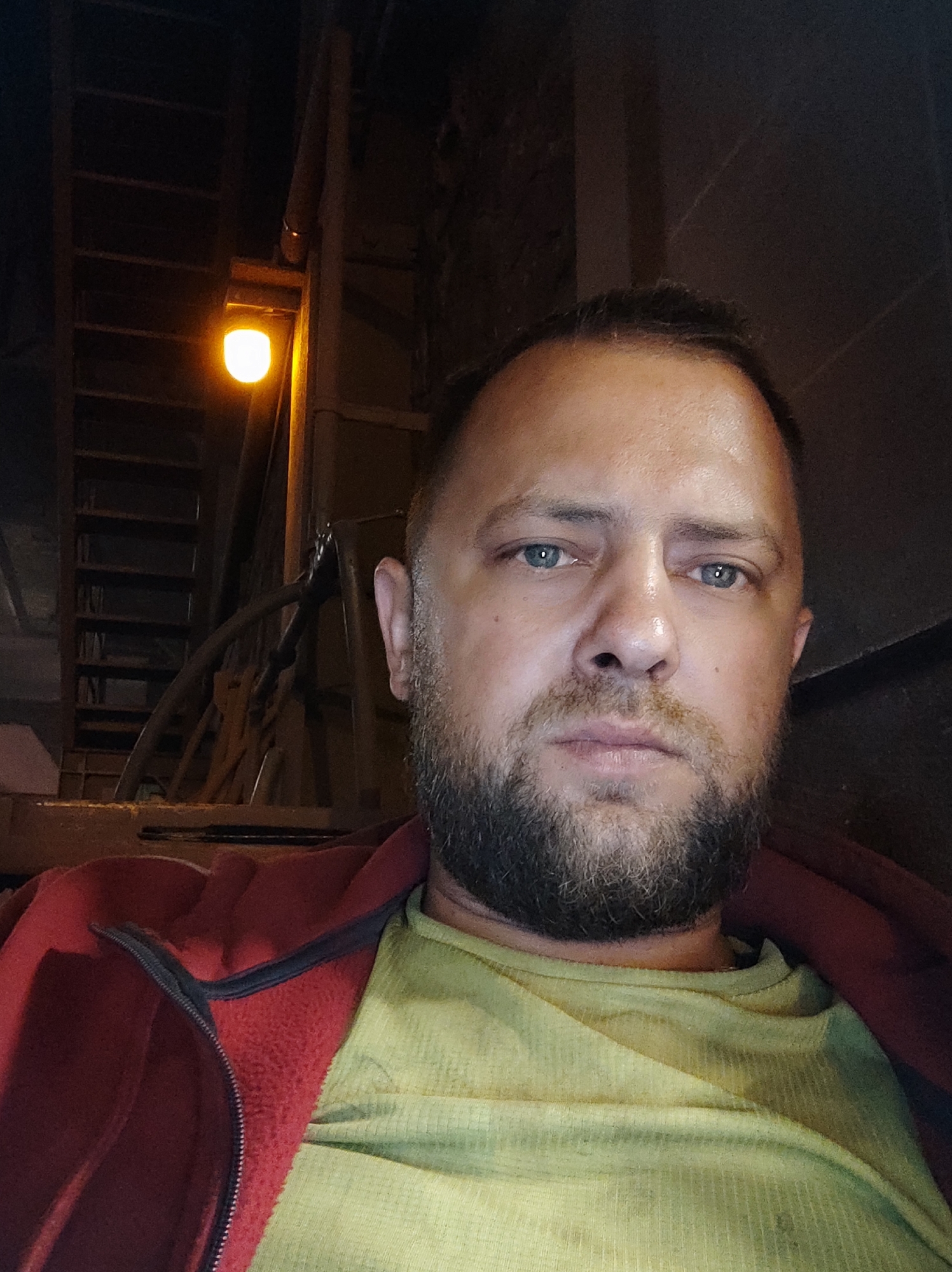A medium shot, selfie style, photo shows a man with a beard and blue eyes, looking directly at the camera. He is wearing a green t-shirt and a red zip-up jacket. The background is dark, with a lit ladder and some industrial-looking structures visible. The man appears to be in an outdoor or semi-outdoor setting, possibly at night, given the lighting. The exact location in Salzgitter, Germany, is not discernible from the image alone.