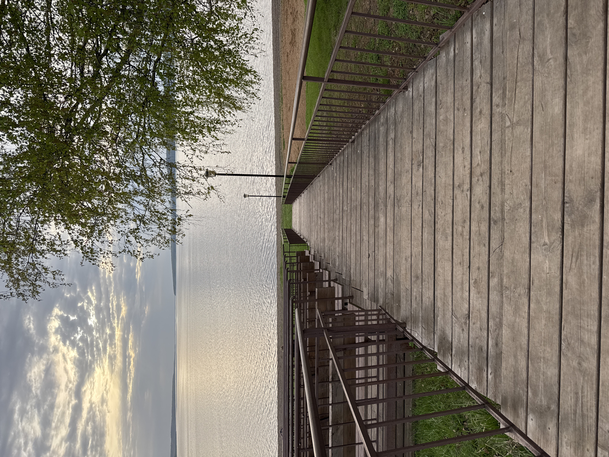 The image depicts a wooden boardwalk with metal railings, likely a pedestrian walkway, leading towards a body of water under a cloudy sky. A tree is visible on the left side of the frame. Two lamp posts stand at a distance along the path. Vegetation (grass) grows between the boardwalk and the railings. The scene may be located in Osa, Russia, given the prompt context. The boardwalk is constructed with parallel wooden planks.