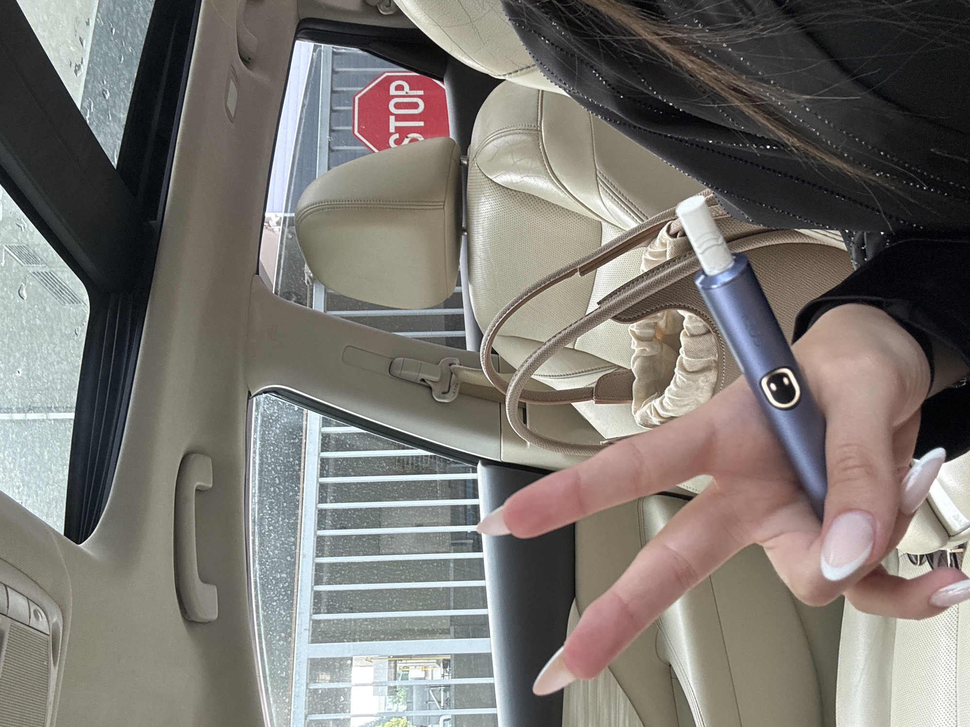 The image depicts the interior of a car, specifically the front passenger area, in Plovdiv, Bulgaria, during or immediately after rainfall, as indicated by visible water droplets on the windows and sunroof. A person, inferred to be female due to manicured hands, is holding a blue electronic device, resembling a heat-not-burn tobacco product, in their left hand while making a V-sign. The hand exhibits long, almond-shaped fingernails with a French manicure. The device features a gold circular button with a stylized face icon and has a white, cigarette-like stick inserted into its top. A portion of the person's torso, wearing a dark garment with vertical lines of subtle embellishments, and long brown hair are visible on the right.
The car's interior consists of light beige perforated leather seats, a matching headliner, and a grab handle above the passenger door. A panoramic sunroof, partially open, is visible, also with raindrops. A light tan handbag with a visible drawstring inner pouch rests on the front passenger seat. Outside the car, through the windows, a white metal fence with vertical bars is prominent. Behind the fence and partially obscured by the front passenger seat's headrest, a red octagonal "STOP" sign is clearly visible. Further in the background, industrial-type structures and green foliage are discernible under overcast lighting conditions.