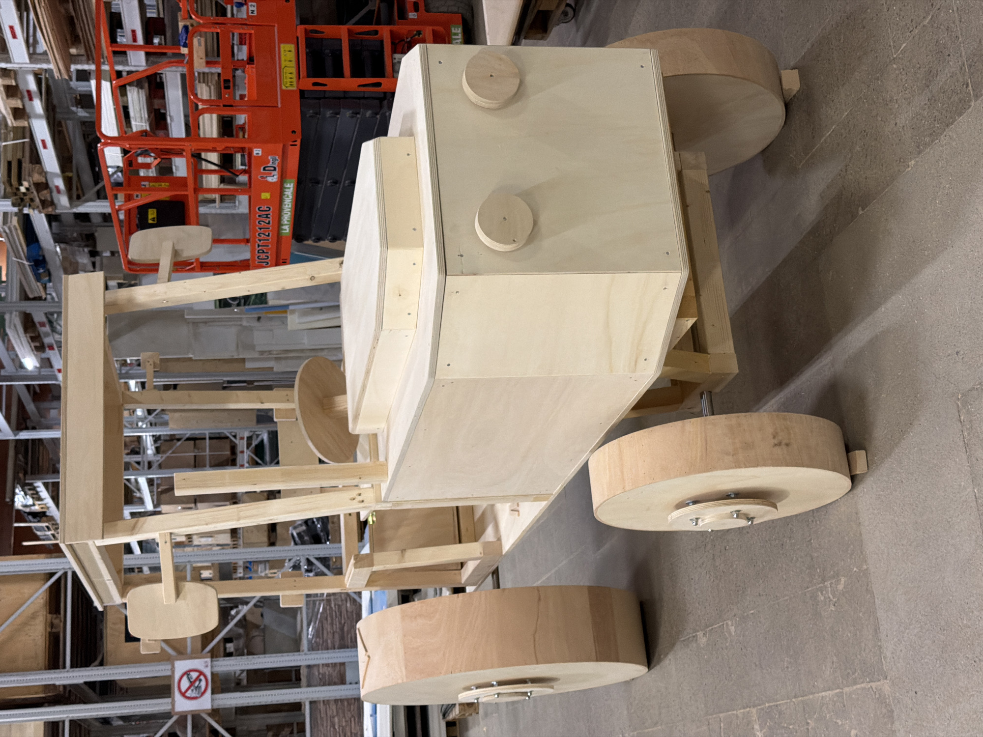 The image shows a wooden model of a tractor under construction, possibly for a school project or event. The tractor is made of unfinished plywood and has large wooden wheels. The construction appears to be taking place indoors, in what looks like a warehouse or workshop. In the background, there are tall shelves stacked with various items, including what appear to be pallets of wood. To the right of the tractor, a bright orange scissor lift is visible, suggesting the space is a storage or industrial facility. The floor is concrete. The overall impression is of an unfinished project in an industrial setting. The specific location of Leudelange, Luxembourg, is not visually discernible from the image itself.