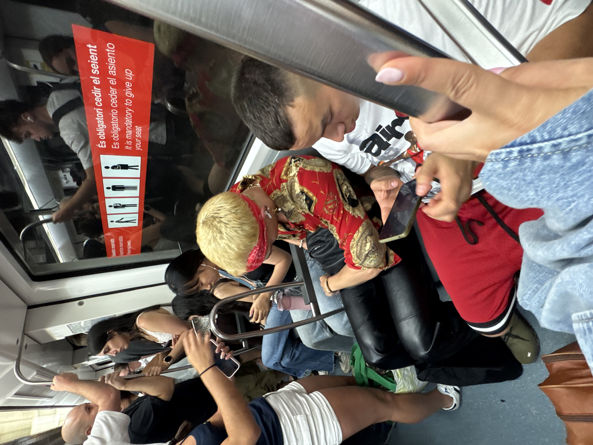 The image depicts the interior of a public transport vehicle, likely a subway or train, with several passengers visible. The environment suggests an urban setting, specifically within the city of L'Hospitalet de Llobregat, Spain, given the multilingual signage visible in the background.
In the foreground, a person wearing white shorts and a dark blue top is standing, their legs and midriff are visible. Next to them, another person is seated, wearing black leather pants and a red shirt with gold and black patterns. This individual also has blond hair and is wearing a red bandana around their head.
Further back, other passengers are seated. One person is wearing blue jeans and a dark top. Another individual, on the right side of the frame, is wearing a white t-shirt with black lettering and is looking down. Their hands are visible, one holding a mobile phone, and another person's hand with light pink painted fingernails is in the extreme right foreground.
The vehicle's interior is characterized by metal poles for support and tinted windows. A red sign with white text in Catalan, Spanish, and English is affixed to the window, indicating a policy regarding seating priority for certain individuals. The flooring appears to be a light blue-grey material.
Several bags are present, including a green shopping bag on the floor and a brown leather bag in the lower left corner. The overall scene captures a moment of daily commute in a populated public transportation setting.