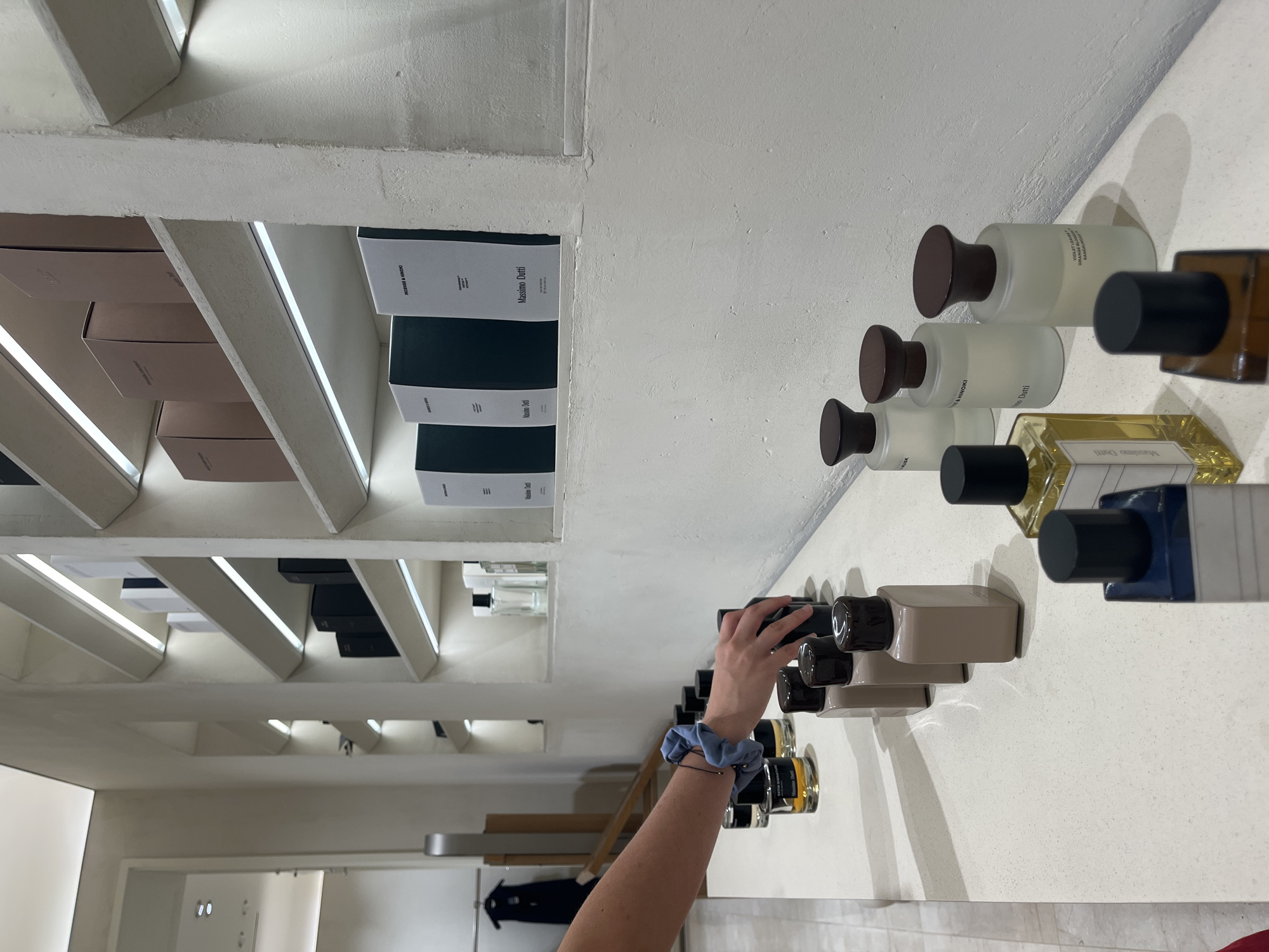 The image shows a person's arm and hand reaching towards a shelf of perfume bottles labeled "Massimo Dutti". The perfumes are on a white shelf, with boxes of Massimo Dutti products stacked on a higher shelf behind. The environment appears to be a store or retail setting. The image is taken from an unusual angle.