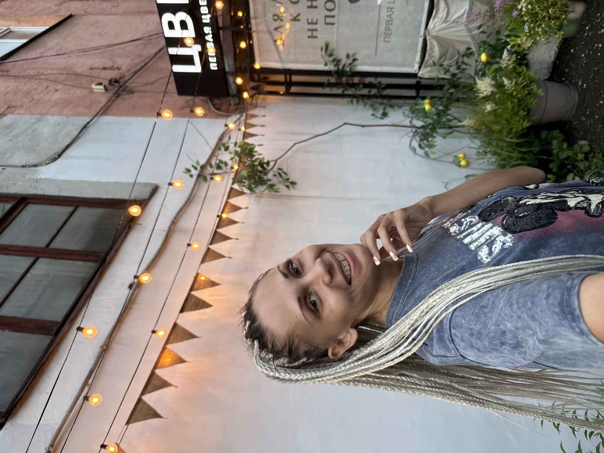 A medium shot captures a young woman with blonde braided hair, facing the camera at a slight angle. She has a warm smile and her right hand is near her chin. She is wearing a grey t-shirt with a graphic print. The background features a white wall adorned with a string of illuminated lights and triangular pennants. To the right, a building with a pink facade is visible, with a sign displaying "ЦВ" in large white letters, followed by smaller text in Russian. Various potted plants, some with yellow decorative elements resembling lemons, are arranged in front of the building. The overall scene suggests an outdoor evening setting, possibly a cafe or shop exterior, located in Fedorino, Russia.