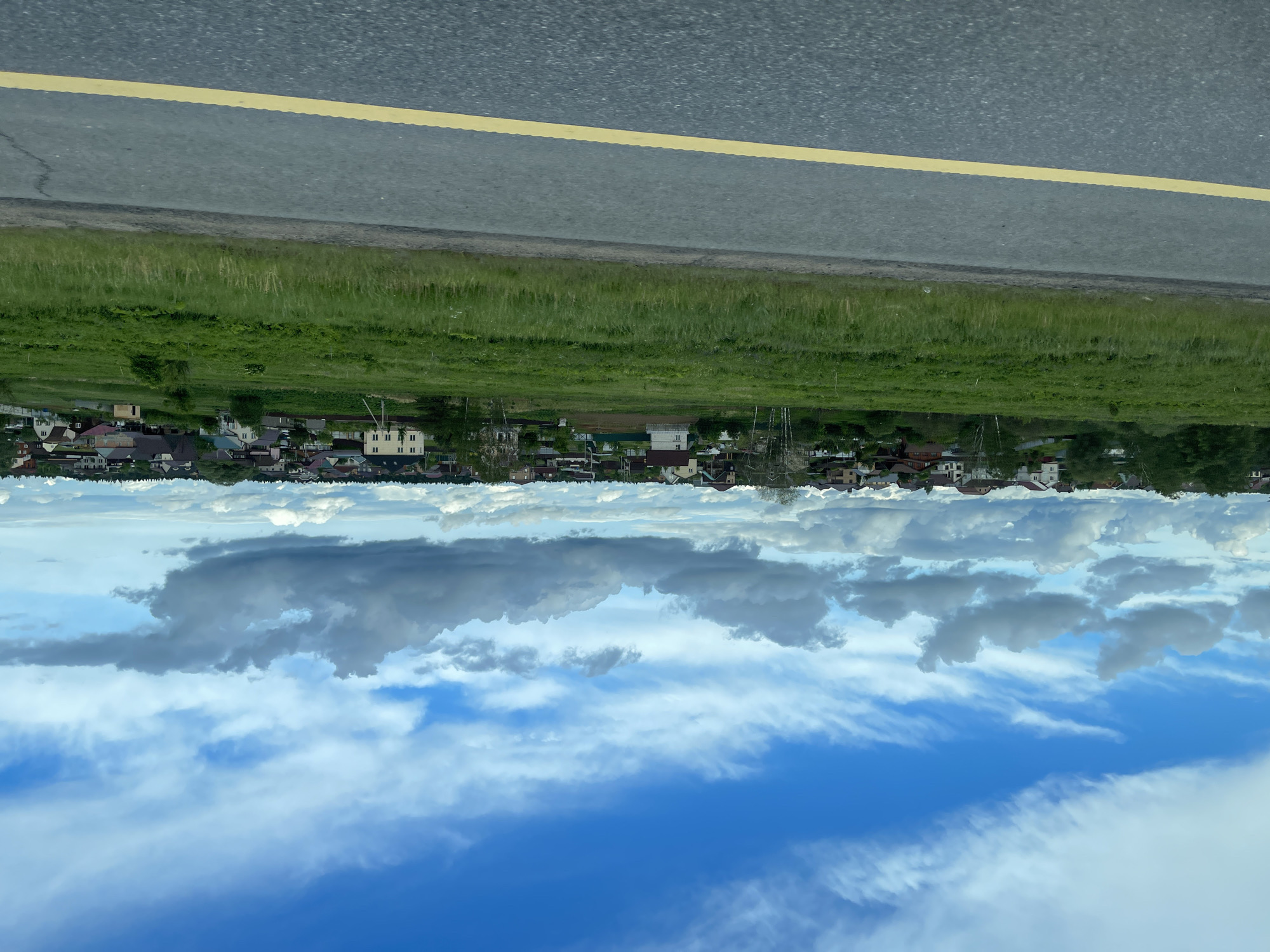 The image depicts a scene in Бавыкино, Russia. It's an upside-down view, showing a blue sky with scattered clouds, a row of houses and trees, a grassy field, and a road with a yellow line. There are no people visible.