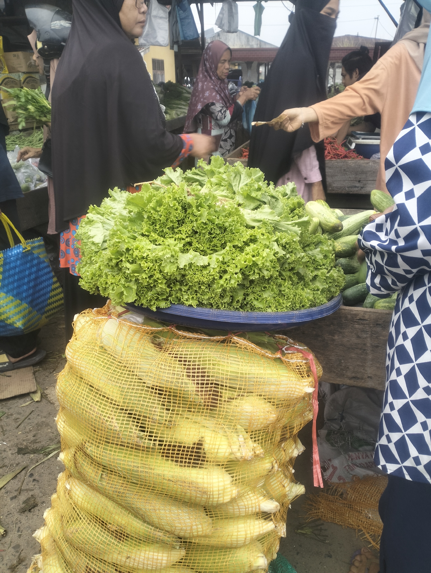 The image depicts a bustling marketplace scene, likely in the city of Dumai, Indonesia. In the foreground, a large sack of corn, its kernels visible through a yellow mesh bag, sits on the ground. Above it, a vibrant mound of leafy green lettuce is displayed in a blue bowl. Several cucumbers are also visible, some in the bowl with the lettuce and others in a wooden crate.
Several individuals are present, engaging in market activities. A person wearing a black hijab is partially visible on the left, facing away from the camera. To their right, another woman in a patterned hijab appears to be interacting with someone off-camera. In the center, a person in an orange garment is handing something, possibly money, to another individual whose face is obscured by a dark veil. A person on the far right is wearing a blue and white patterned outfit.
The environment suggests an outdoor market with wooden stalls and a somewhat dusty ground. The background shows additional stalls and structures, hinting at the lively commercial atmosphere. The overall impression is one of a typical Indonesian wet market, characterized by fresh produce and active commerce.