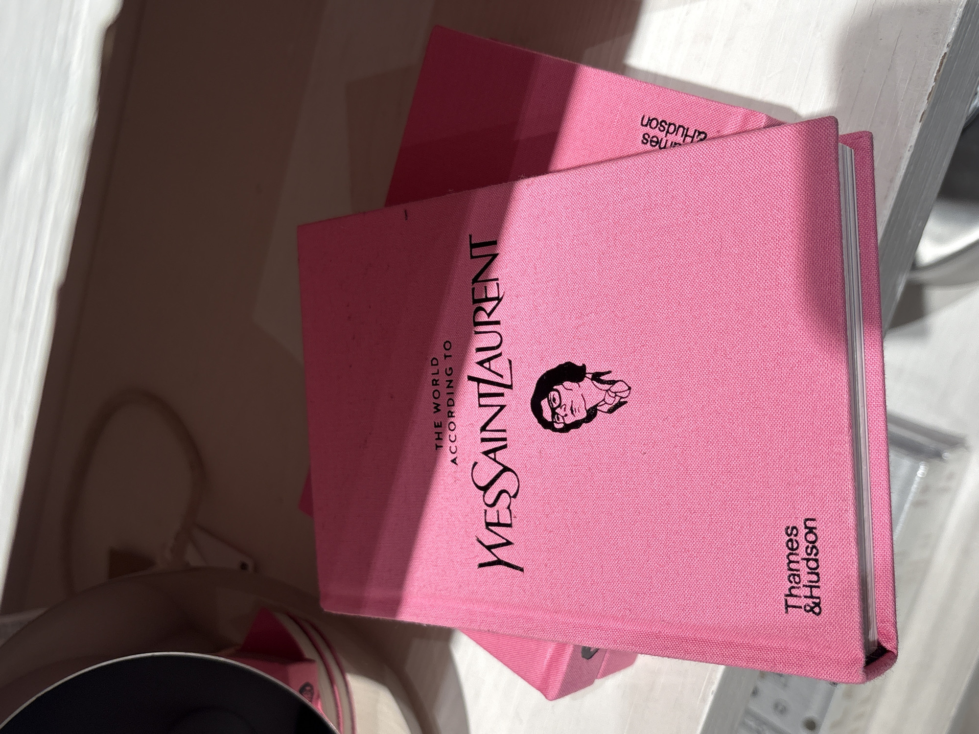 Here's a detailed description of the image:
The image shows two pink books. The top book has the title "YVES SAINT LAURENT: THE WORLD ACCORDING TO." It features a black silhouette illustration and the publisher name "Thames & Hudson." The second book appears to be a similar pink color, partially covered by the top book. The books appear to be on a surface. The background includes a portion of a dark-colored item, and to the right, a glimpse of a white surface. There are no people visible in the image. The photo appears to be indoors under somewhat bright lighting, casting a shadow across the scene.
This image does not contain any information which would allow the determination of the location of the photo being taken in Sofia, Bulgaria, or any other location.