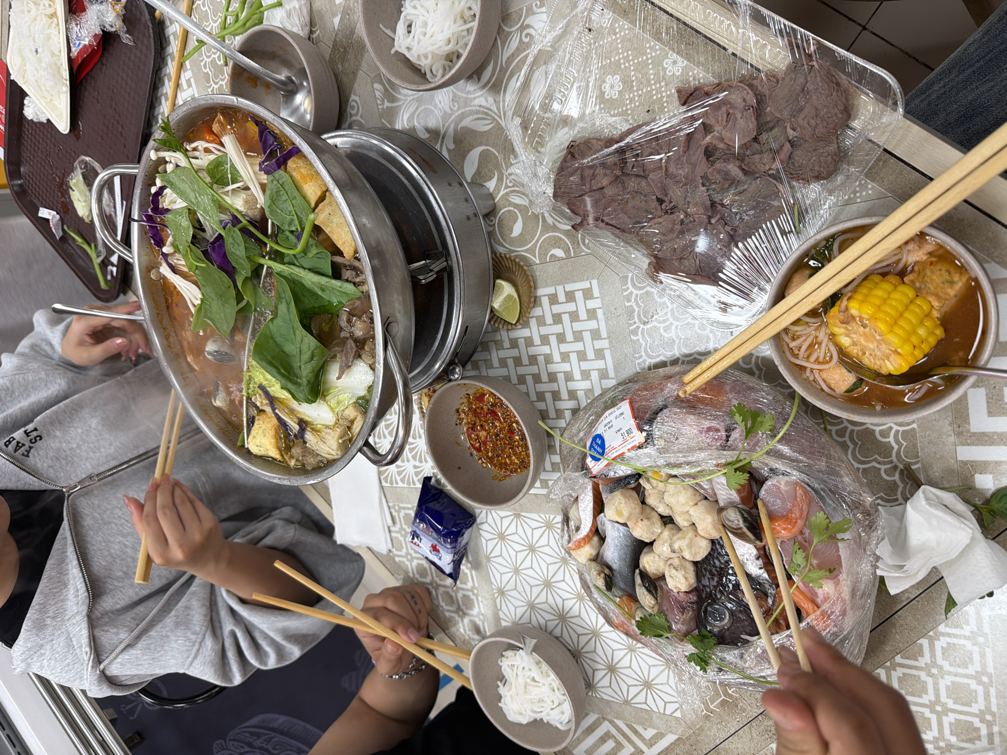 An overhead shot captures a lively hot pot meal shared by at least three individuals at a table, likely indoors. The central focus is a large, steaming hot pot filled with a rich broth, various vegetables including spinach, mushrooms, and red cabbage, along with tofu and what appears to be pre-cooked meat.
Around the hot pot, several components for the meal are arranged. A platter of thinly sliced raw beef, covered in plastic wrap, awaits cooking. Adjacent to it, a large, transparent plastic-wrapped package holds an assortment of fresh seafood, including pieces of fish (identifiable as tilapia from the visible label "CÁ DIÊU HỒNG"), shrimp, clams, and fish balls, garnished with fresh cilantro. A white sticker on this package clearly shows "SIÊU THỊ" (supermarket), "ĐÀ NẴNG" (Da Nang), and a price of "51.900 Đ".
Diners use chopsticks, seen in multiple hands, to pluck ingredients from the hot pot or to serve themselves. Small bowls of white vermicelli noodles and a bowl of chili flakes or sauce are also on the patterned table. One person is wearing a grey zipped hoodie with "FAB ST" visible on the sleeve. A small bowl containing hot pot soup, corn on the cob, and noodles is visible, suggesting individual servings are being enjoyed. A lime wedge sits on a woven coaster, and a blue packet of seasoning or condiment is also present. The scene conveys a communal and casual dining experience, typical of a hot pot meal in Vietnam.
