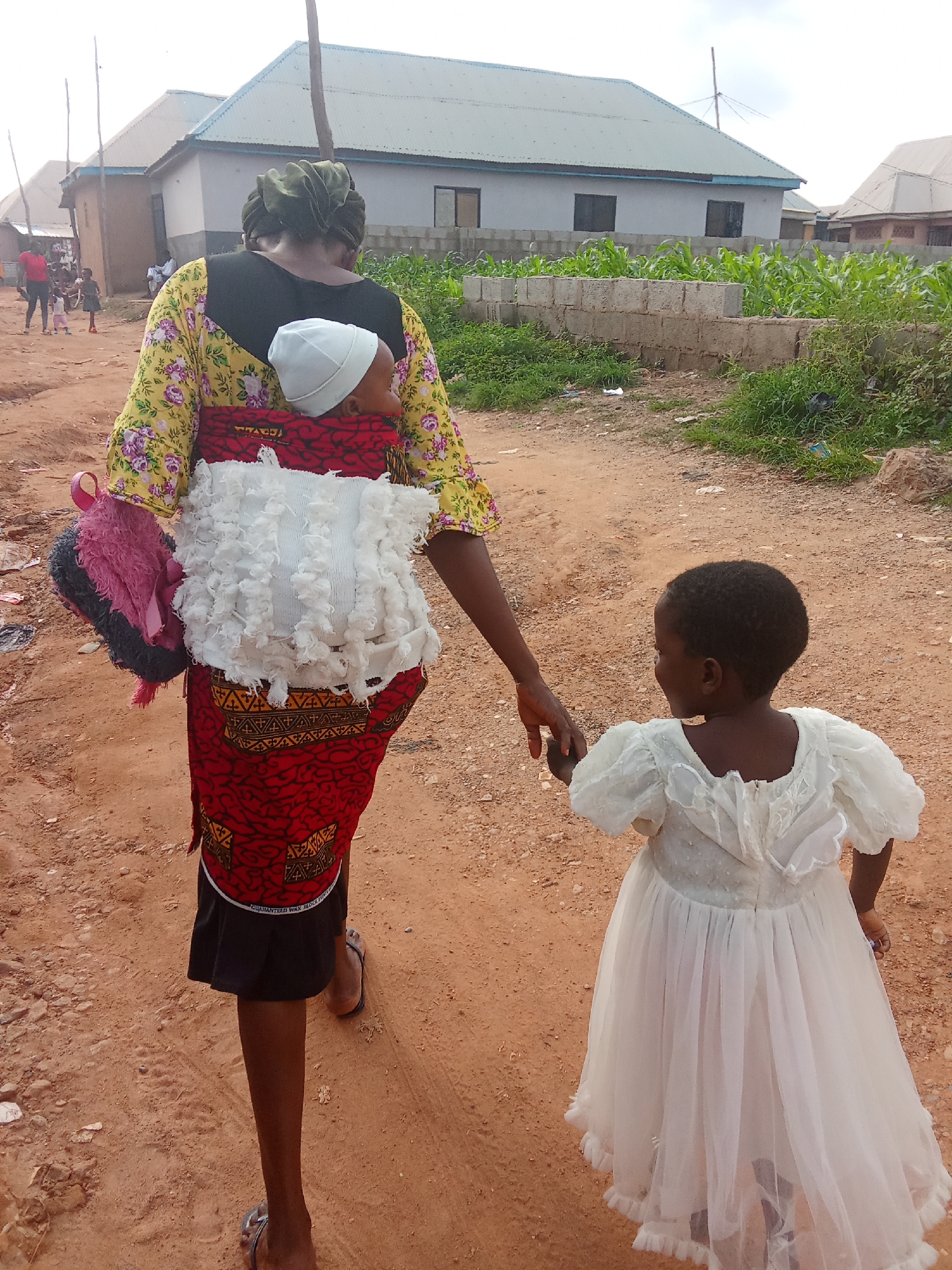 In Kagini, Nigeria, a woman walks on a dirt road carrying a baby on her back. The baby is wrapped in a white cloth with ruffles and a red and black patterned fabric is visible around the baby. The woman wears a yellow and floral print top and a red and black patterned skirt. She is holding the hand of a young girl in a white dress with a full skirt. In the background, there are houses with gray roofs and a plot of land with green plants, likely corn, bordered by a concrete block wall. The ground is covered in reddish-brown dirt and small stones.
