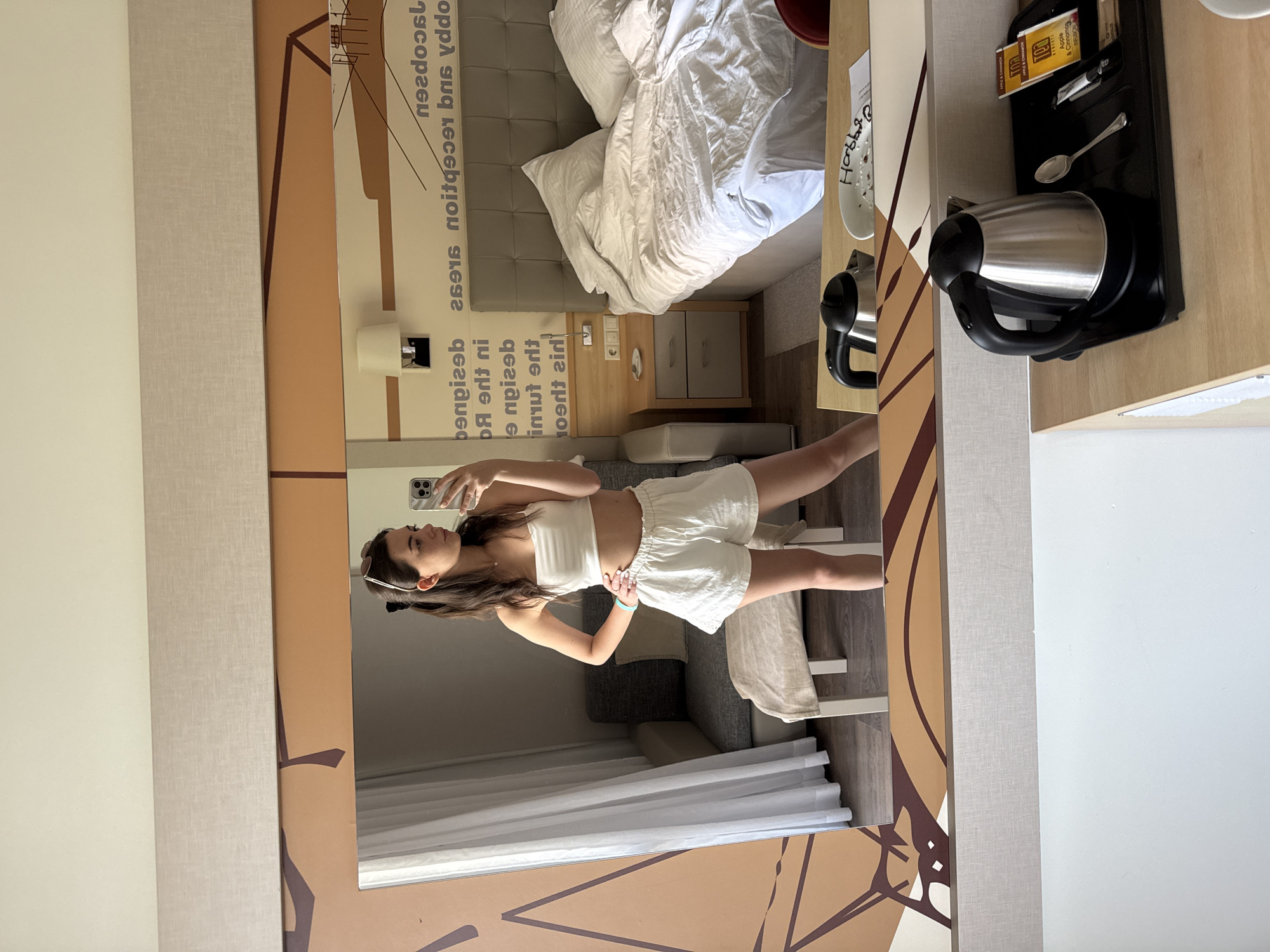 A young woman with dark hair pulled back in a ponytail, wearing a white strapless top and white linen shorts, poses for a mirror selfie in a hotel room. She is standing with one hand on her hip and the other holding a smartphone. The room features a large bed with rumpled white bedding, a nightstand with a lamp, and a small seating area with a couch and ottoman. A mirror dominates the foreground, reflecting the woman and the room. A kettle, spoon, and tea packets are visible on a tray on a wooden surface in the lower part of the frame. A decorative wall graphic with brown and tan geometric patterns is visible on the wall behind the mirror.