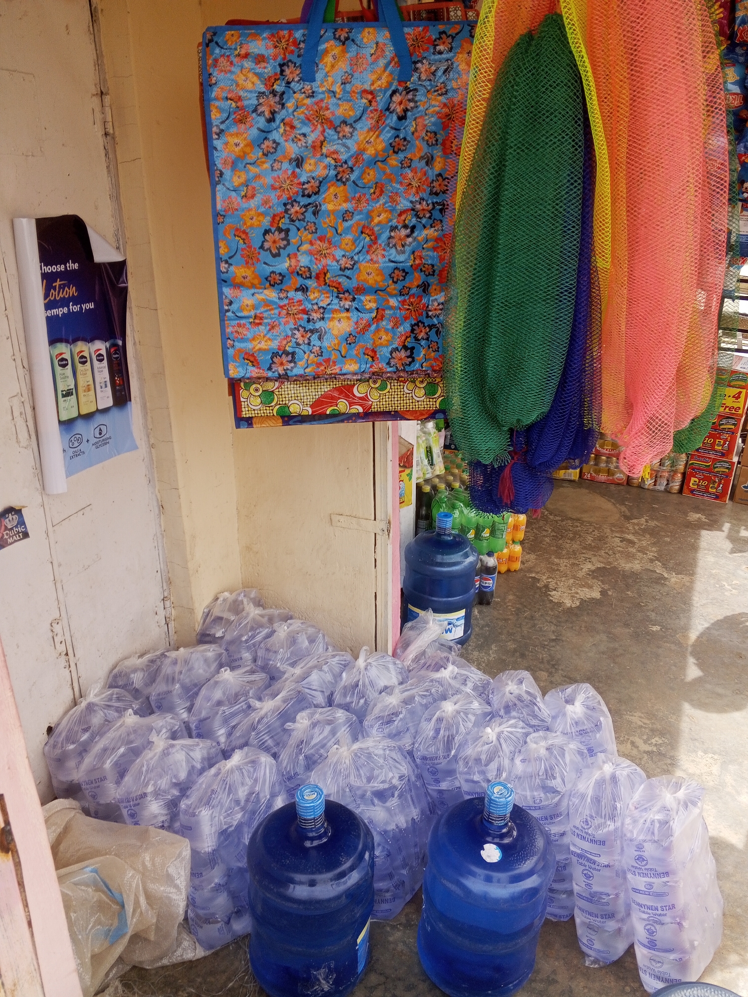 The image captures a street-side commercial scene. Stacks of individual water bags labeled "BENNYNEN STAR Table Water" are grouped together on the ground. There are also three large, blue, refillable water jugs. Above the jugs are hanging colorful mesh bags. A display for body lotion is seen on the wall, next to the water. The scene seems to be an informal retail setup in an area where "BENNYNEN STAR Table Water" is commonly sold. Other items can be seen on the wall in the upper right side of the image.