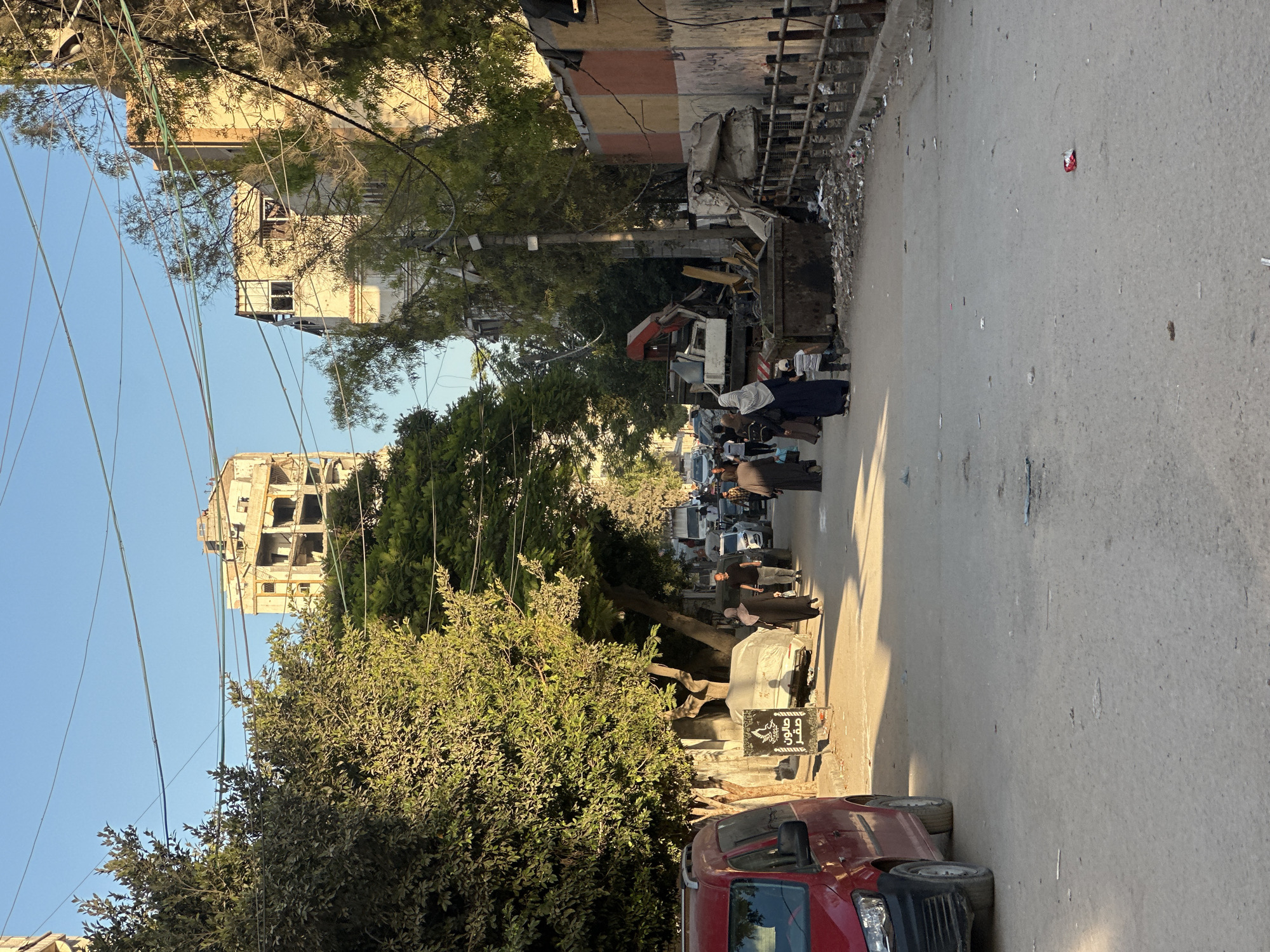 In a city street under a clear blue sky, a red van is parked on the left, facing right. Beyond it, a street lined with trees leads into the distance. Several people are walking on the sidewalk on the left side of the street, and on the right side, there are buildings under construction or in disrepair, a dumpster filled with debris, and a construction vehicle. The street is paved and appears somewhat worn, with some litter scattered on the ground. The overall scene suggests an urban environment with ongoing construction or demolition work.