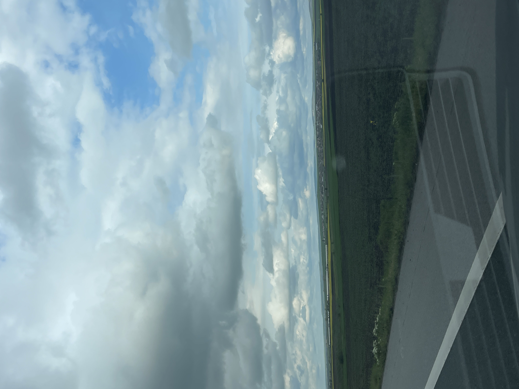The image, originally rotated 90 degrees clockwise, captures a rural landscape viewed from a vehicle, likely in Vodenichane, Bulgaria, during daytime. The upper two-thirds of the corrected orientation show a vast sky filled with large, fluffy cumulus clouds in shades of white and light grey, interspersed with clear patches of vibrant blue sky.
Below the horizon, the landscape unfolds with extensive green agricultural fields, suggesting a fertile plain. A distinct band of yellow fields, possibly rapeseed, can be seen running horizontally across the mid-distance. Further in the background, a faint line of buildings or a small settlement is discernible against the distant horizon.
The lower right portion of the image is dominated by the reflection of the vehicle's interior on the window. This reflection includes a grey, ribbed structure, possibly an air vent or part of the vehicle's console, and a dark, textured panel. A thick, light-colored line, potentially a road marking or another vehicle component, is also visible within this reflection. The window surface shows some dust or smudges.
The weather appears partially cloudy, with soft, diffused light illuminating the scene. No people are visible, and there is no discernible text in the image. The overall impression is a calm, observational view of a peaceful countryside.