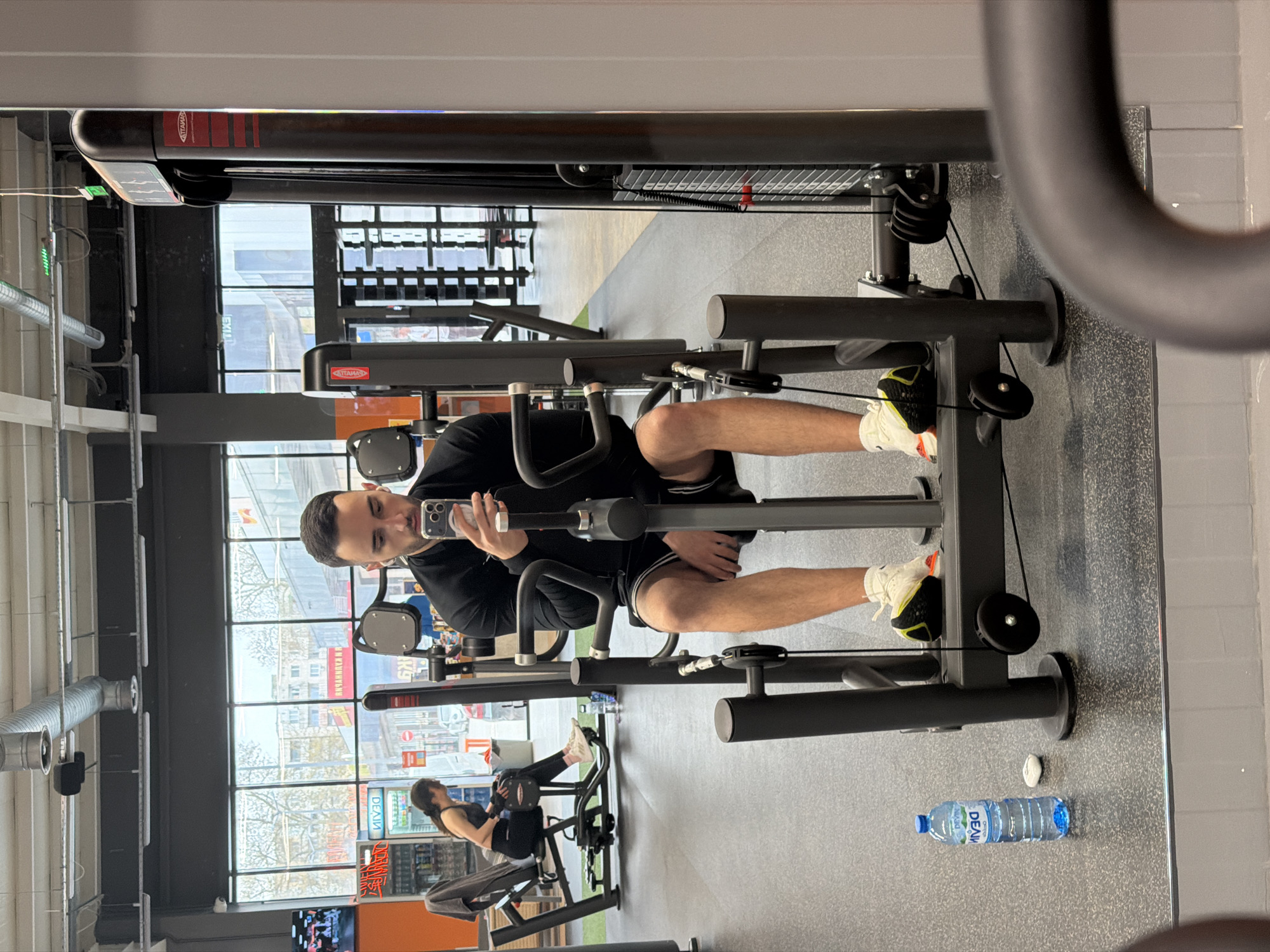 The image depicts the interior of a gym. A man in a black shirt and shorts is sitting on a leg extension machine, taking a selfie with his phone. The gym equipment is black and silver. A woman is using another piece of gym equipment in the background. A water bottle is visible on the floor next to the man. The background features large windows that reveal a view of the city. Red text visible in the windows reads "DELIA". Based on the "DELIA" signage and the architectural style visible through the windows, the location is most likely in Plovdiv, Bulgaria.