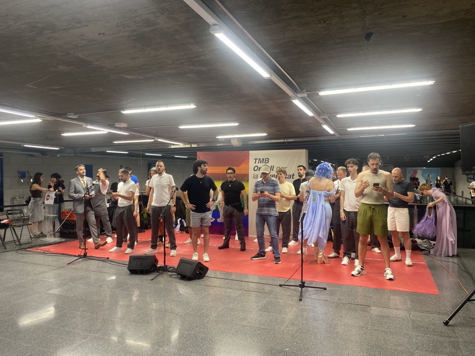 The image captures a gathering of approximately fifteen people on a red carpeted platform, possibly a makeshift stage, under a concrete ceiling with exposed lighting fixtures. The setting appears to be an indoor public space, potentially a transit station given the industrial aesthetic of the ceiling and the presence of what looks like a banner with text in Catalan, suggesting Barcelona, Spain.
In the foreground, several men are standing. Some are holding microphones or appear to be about to speak. One man in the center is wearing a light blue, flowing dress and has blue hair. Another man is holding a phone, looking down at it. To the left, a man in a suit and vest is addressing the group. In the background, other individuals are observing, including women wearing casual attire.
Objects in the scene include microphones on stands, speakers, a podium, chairs to the left, and bags or equipment on the ground. The overall atmosphere seems to be that of a performance, presentation, or public event. The banner behind the central group reads "TMB" followed by "Orgull per la Llibertat" which translates to "Pride for Freedom," indicating a Pride-related event.