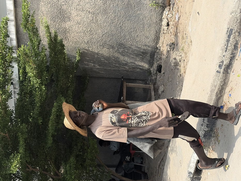 The image captures a street scene in Igboefon, Nigeria, under daylight conditions. A dark-skinned adult male occupies the central foreground, walking from left to right on a light-colored, slightly elevated pathway. He wears a wide-brimmed straw hat, a light brown t-shirt with a graphic print depicting a masked face featuring red accents, dark trousers with a light vertical stripe on the left leg, and dark flip-flops. A grey, possibly cloth, bundle or bag is slung over his left shoulder, and he holds a small, dark circular object with a yellow ring in his right hand. A thin white wire is visible near his right ear. To the left of the main subject, under the dense foliage of a large green tree, two other individuals are partially visible; one is seated, wearing a light blue shirt, and appears to be using a mobile phone. A crude wooden table is also present in this shaded area. The background to the right consists of a textured, light grey wall of a building. The ground immediately adjacent to the pathway is unpaved, dusty, and contains scattered debris and sparse, small plant growth. Shadows suggest a sunny environment.